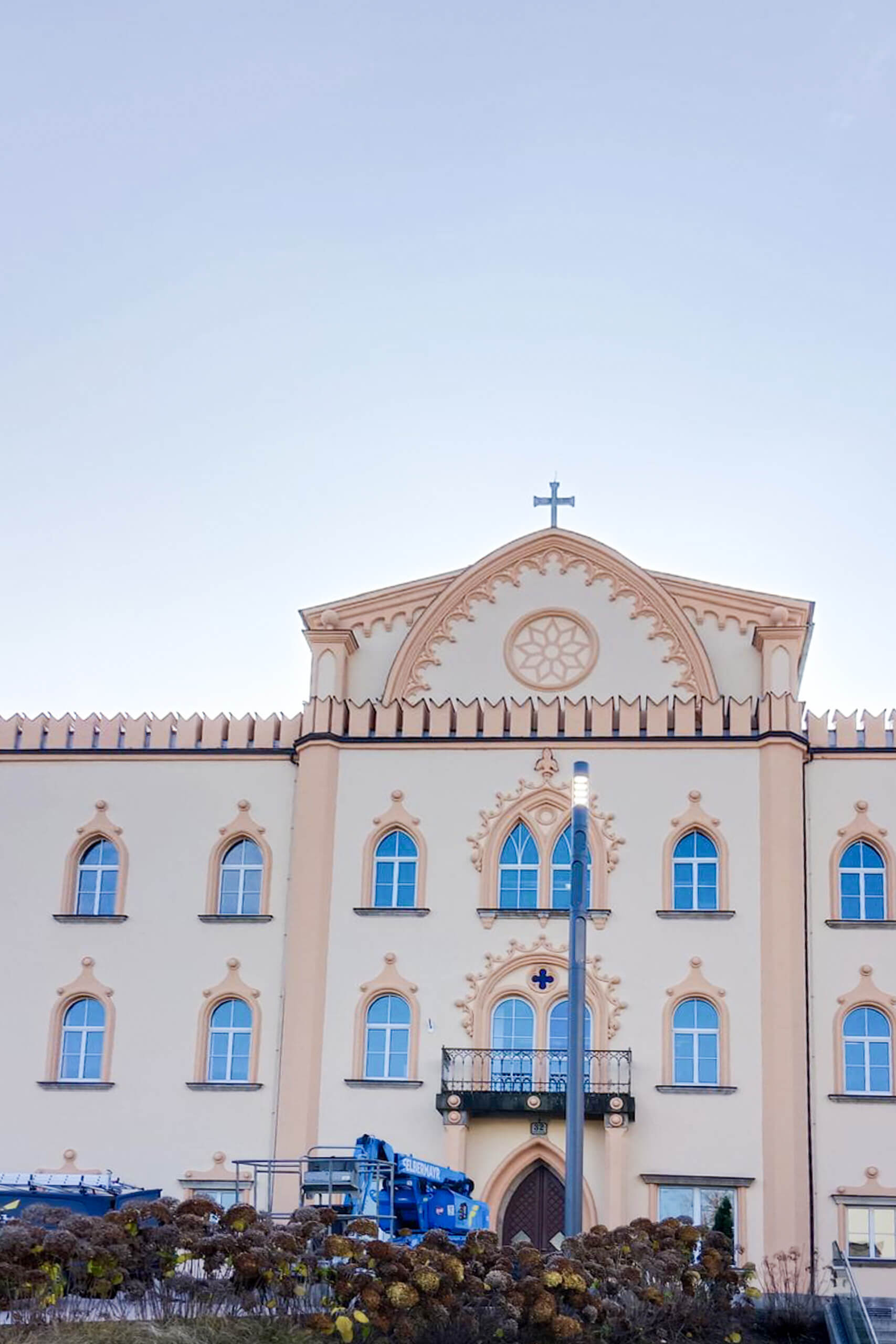 Fassade eines Gebäudes mit Kreuz und Rundbogenfenstern, beleuchtet durch eine moderne Straßenlaterne.