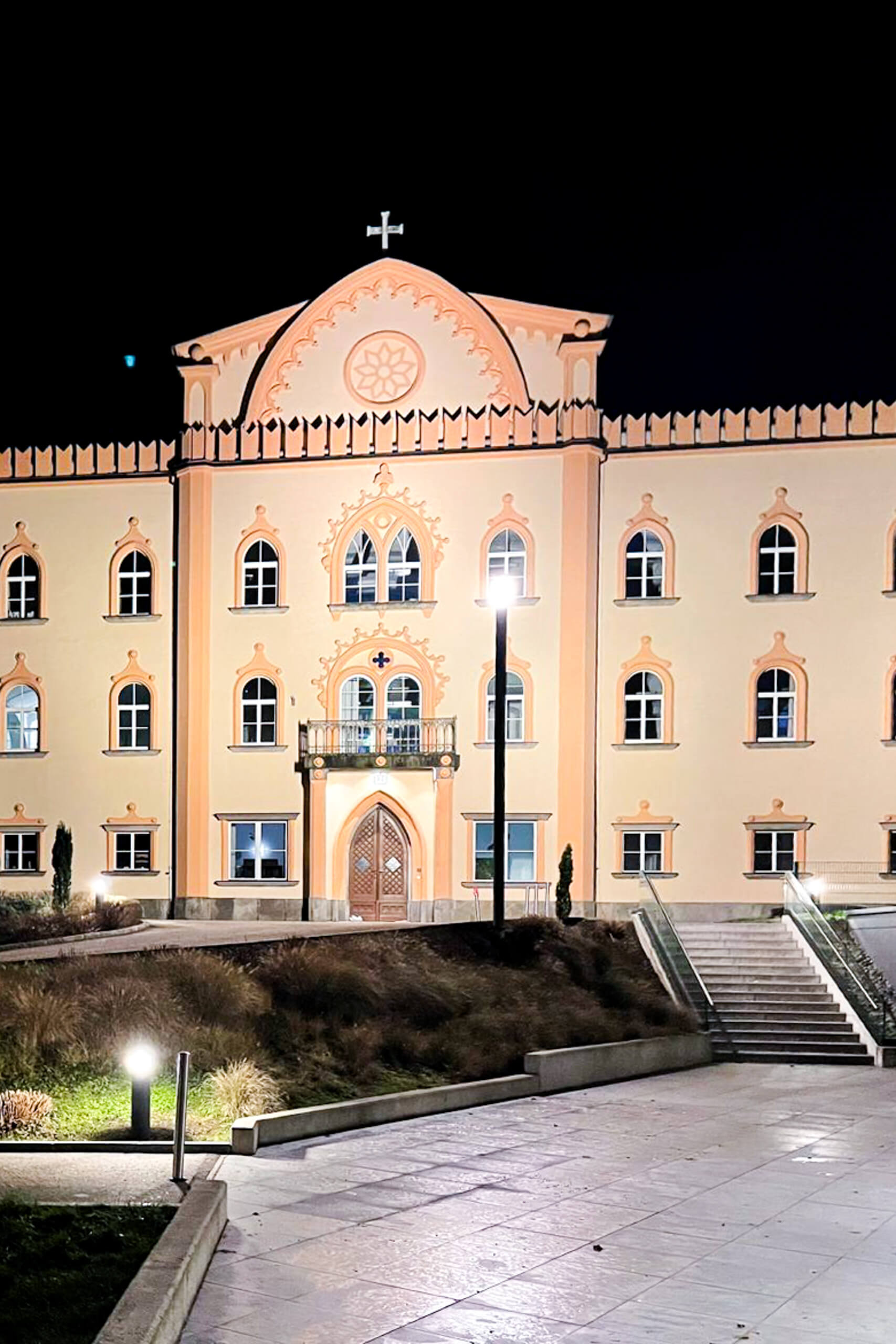 Beleuchtetes Klostergebäude bei Nacht mit Treppenaufgang und Parkanlage