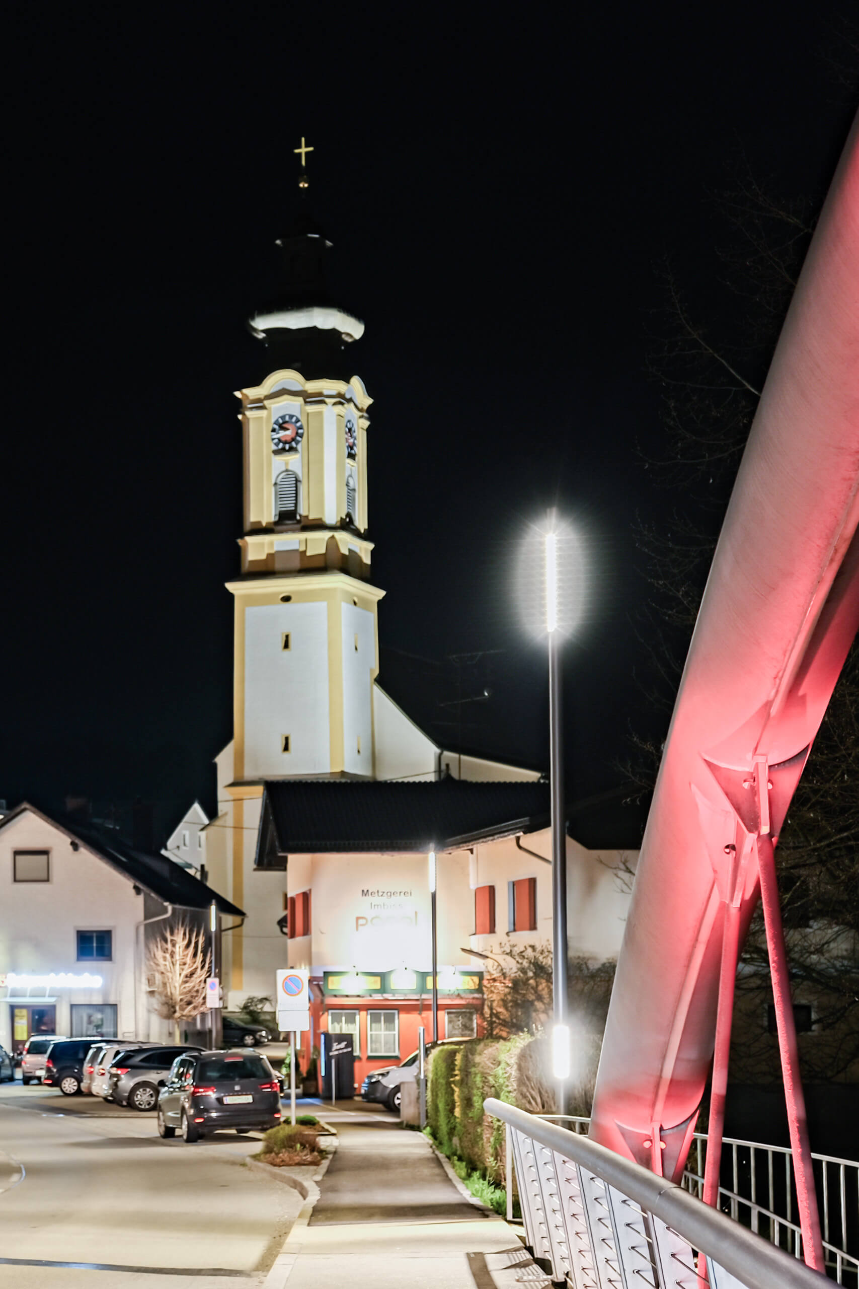 Nachtaufnahme einer beleuchteten Kirche und eines Metzgereiladens mit Straßenlaternen.