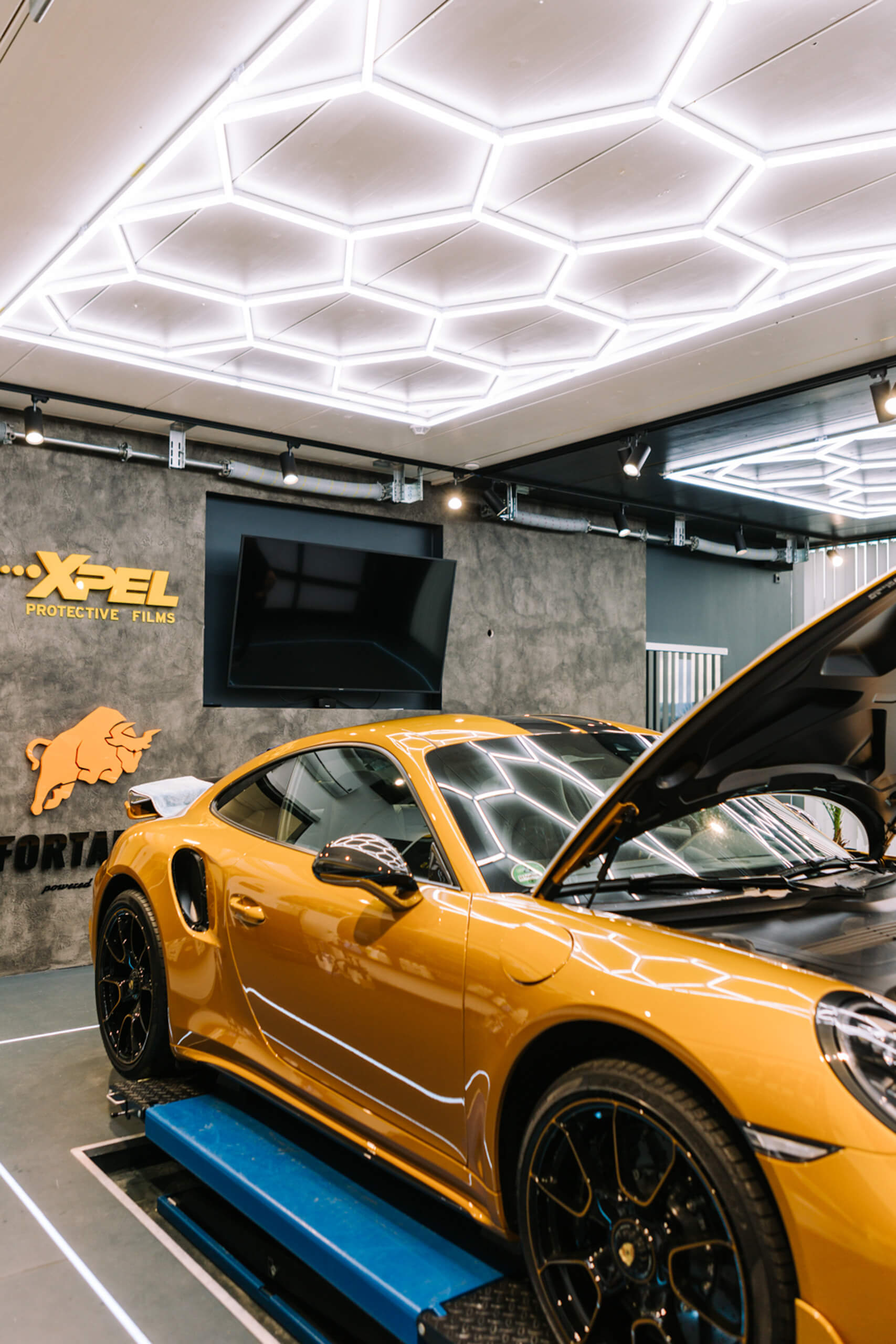 Goldener Porsche 911 mit geöffneter Motorhaube in einer Werkstatt mit hexagonalen Deckenleuchten