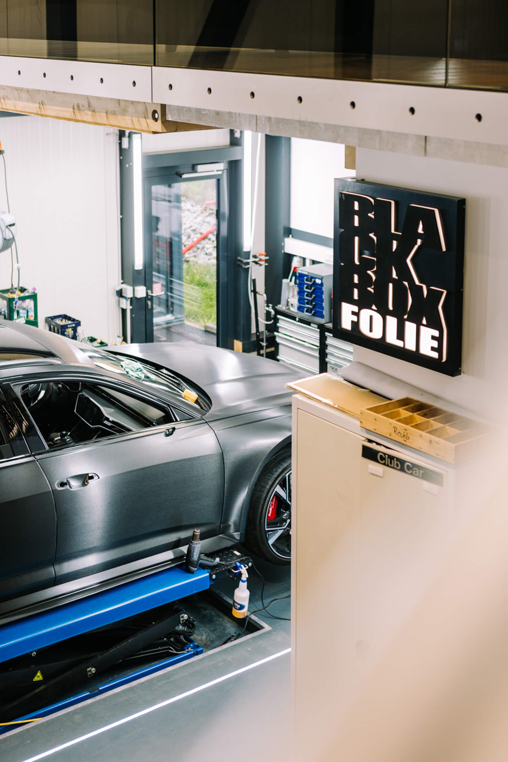Graues Auto auf Hebebühne in Werkstatt mit Schild "BLACKBOX FOLIE