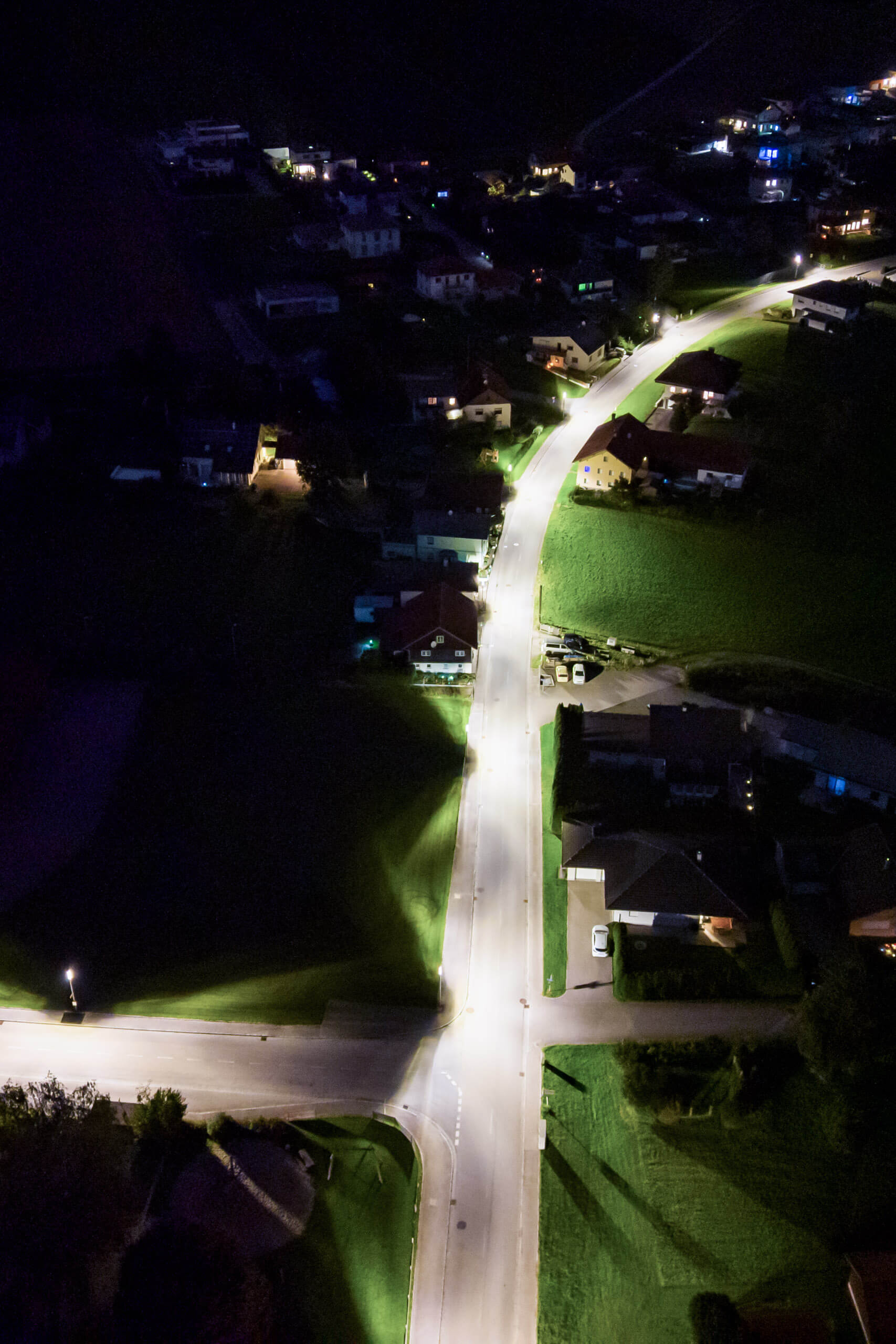 Luftaufnahme einer beleuchteten Straße in einem Dorf bei Nacht mit Häusern und Grünflächen.