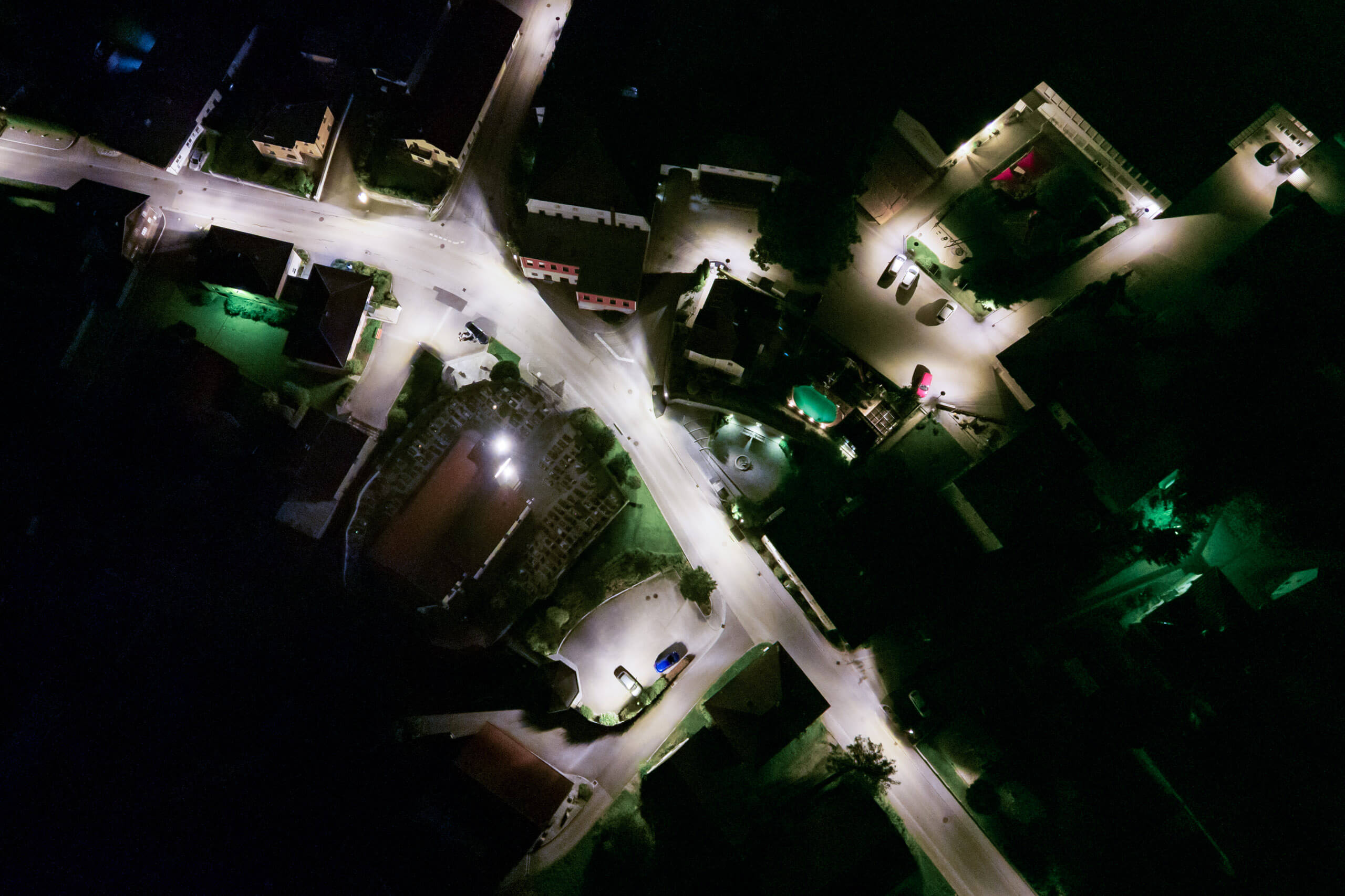 Luftaufnahme eines beleuchteten Dorfplatzes bei Nacht mit Straßenlaternen und Gebäudefassaden
