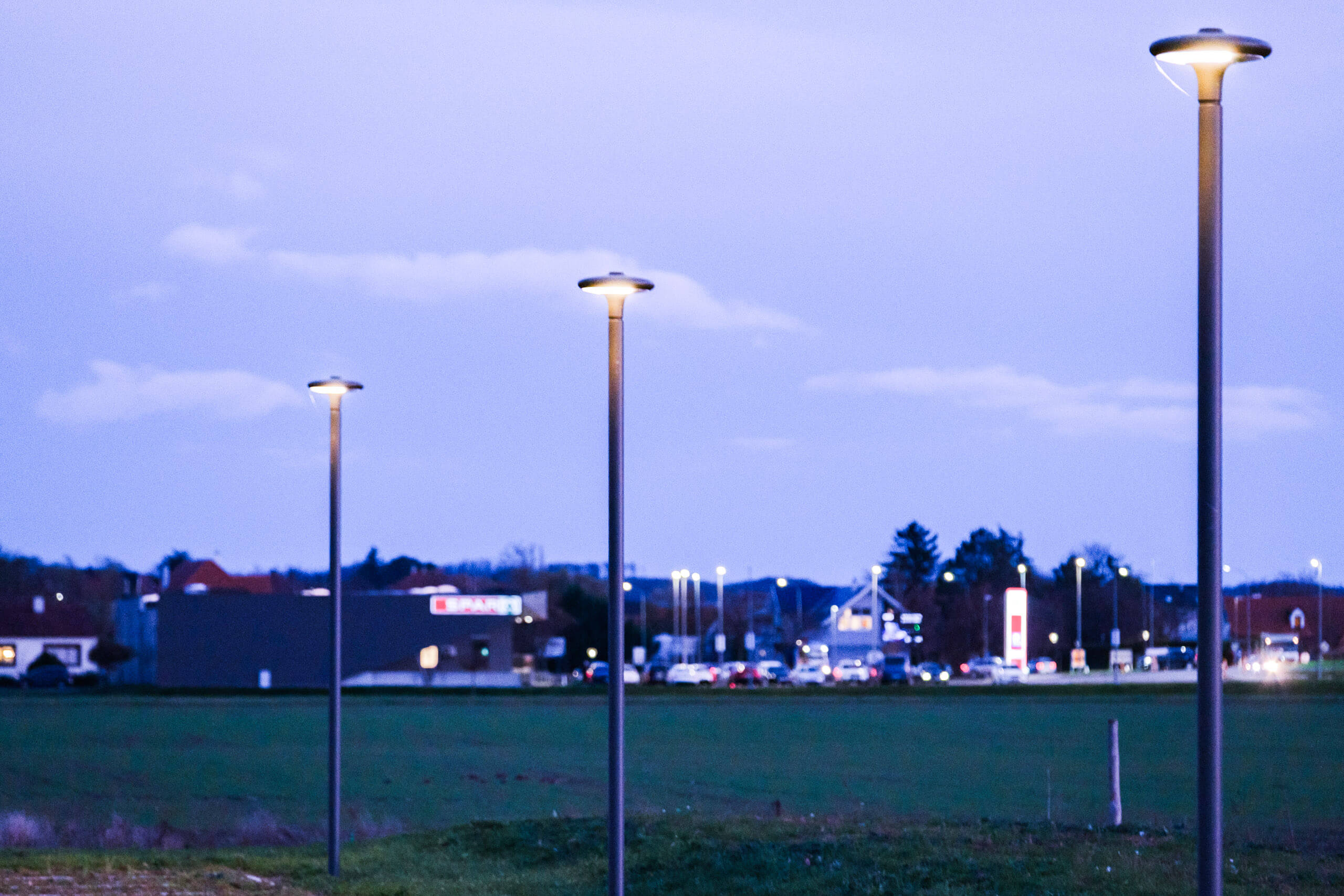 Drei moderne Straßenlaternen im Abendlicht vor einer Siedlung mit Geschäften.