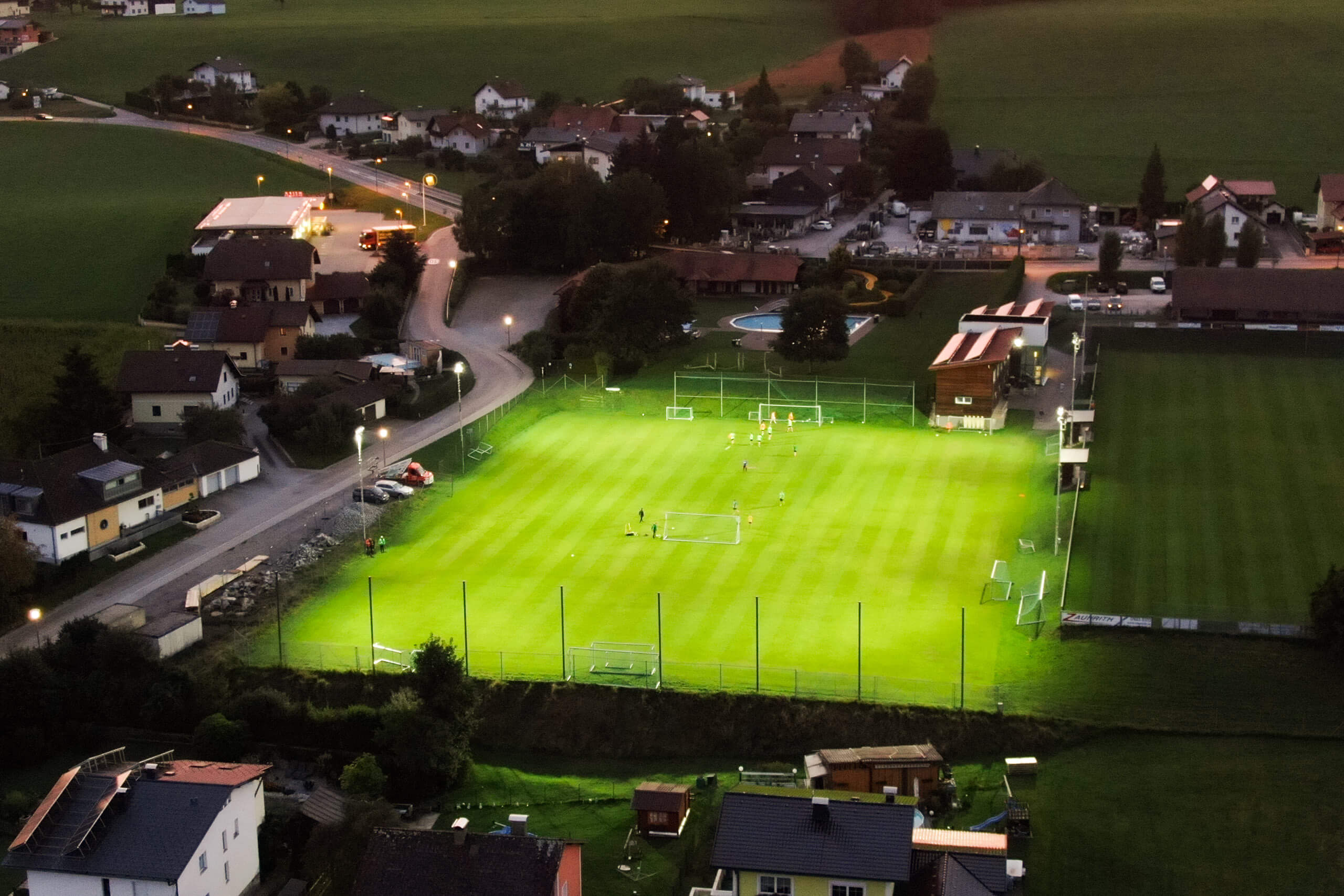 Beleuchteter Fußballplatz bei Dämmerung mit Spielern und umliegenden Häusern.