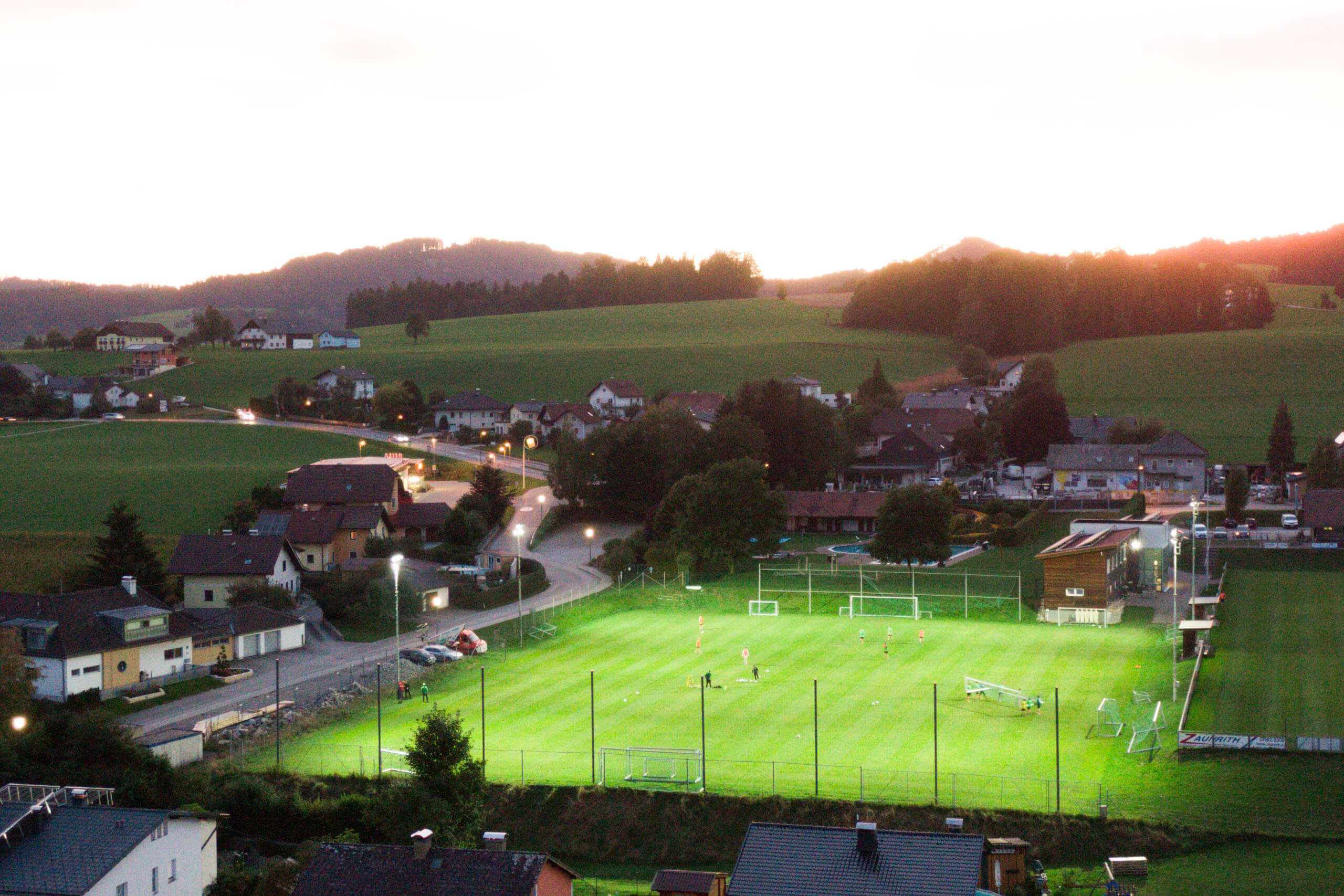 Beleuchtetes Fußballfeld mit umliegender Ortschaft bei Dämmerung, Sportplatzbeleuchtung von Illumina