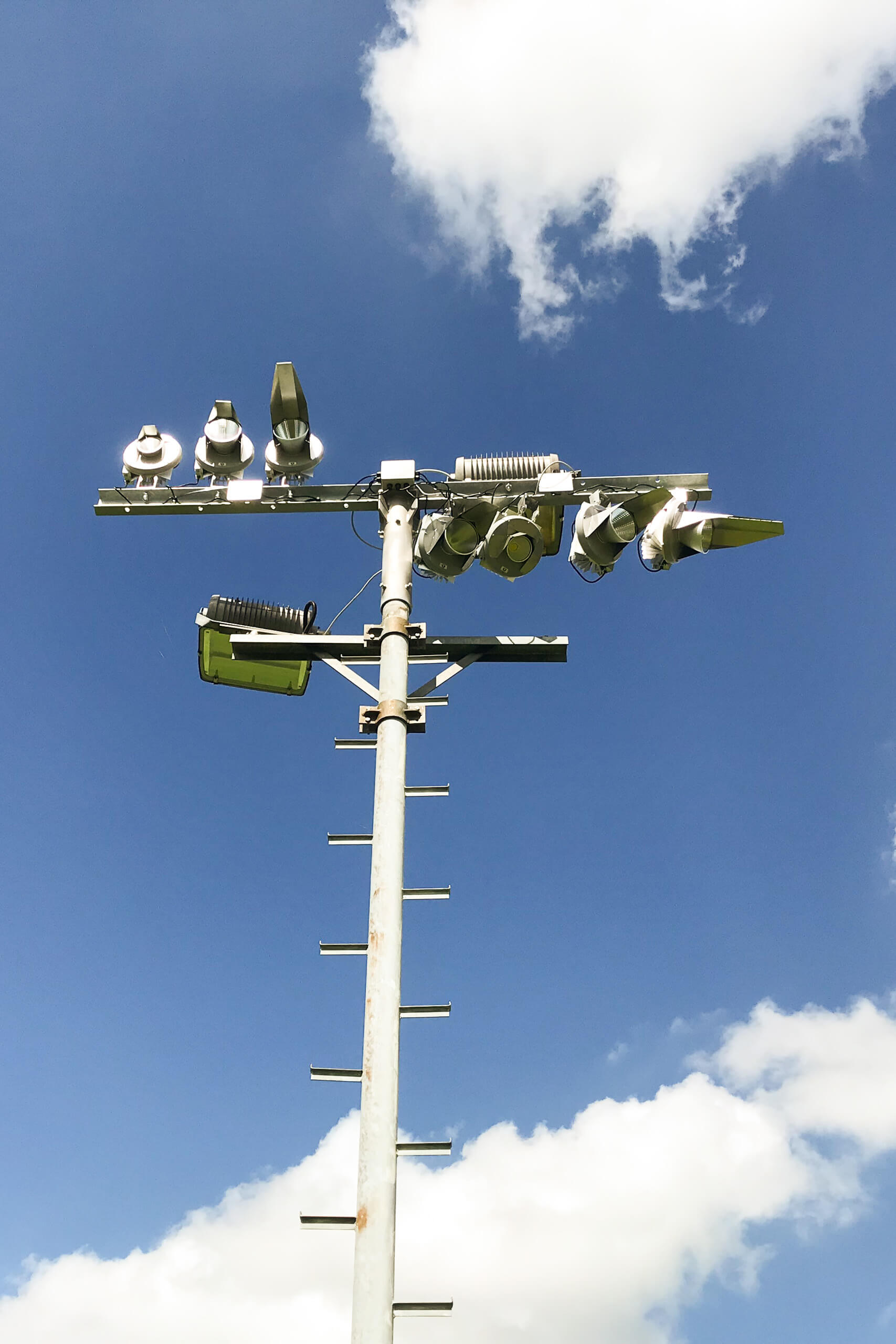 Beleuchtungsmast mit Flutlichtscheinwerfern und Leiter gegen blauen Himmel mit Wolken.