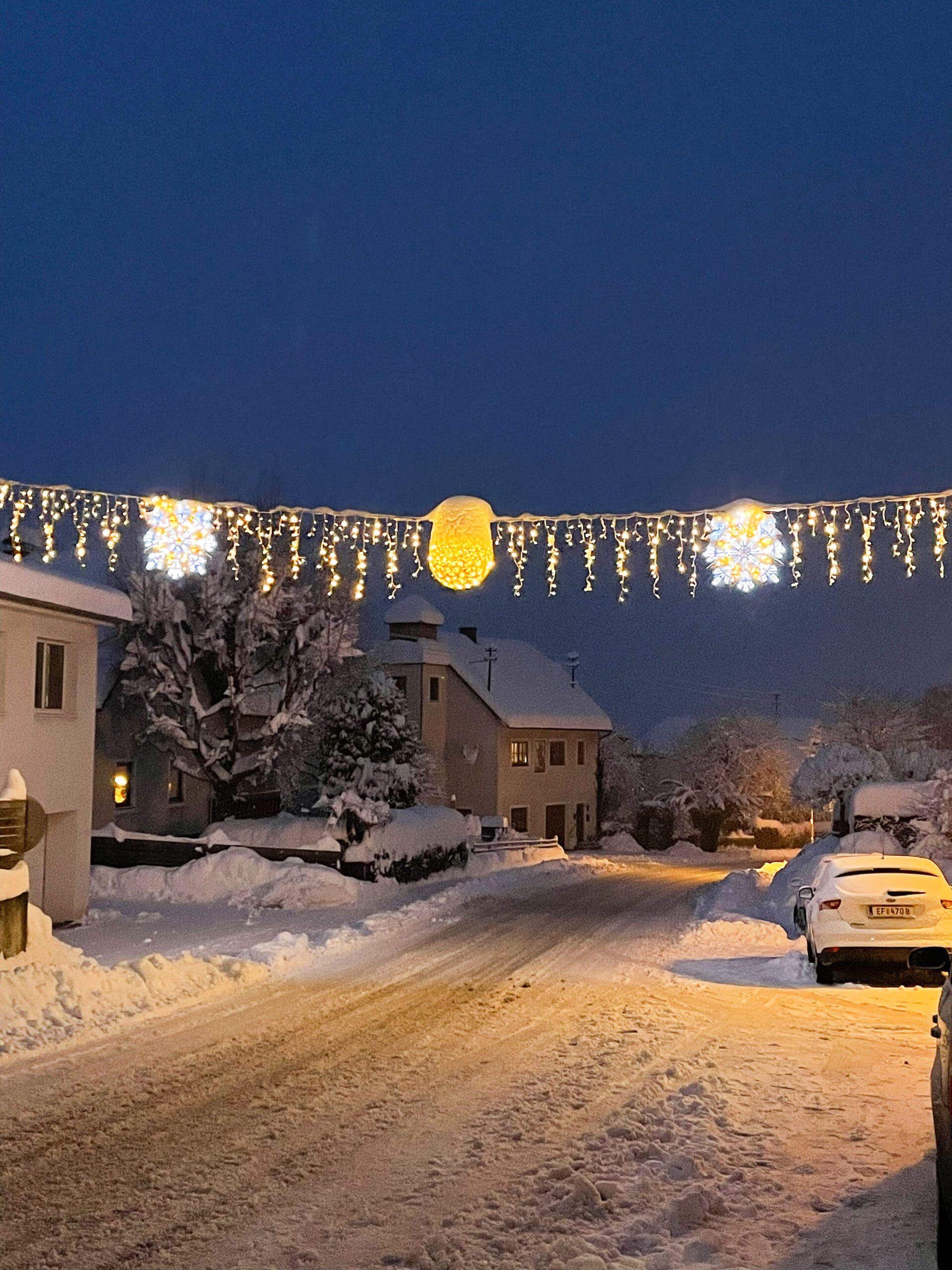 Schneebedeckte Straße mit Weihnachtsbeleuchtung und Häusern im Abendlicht.