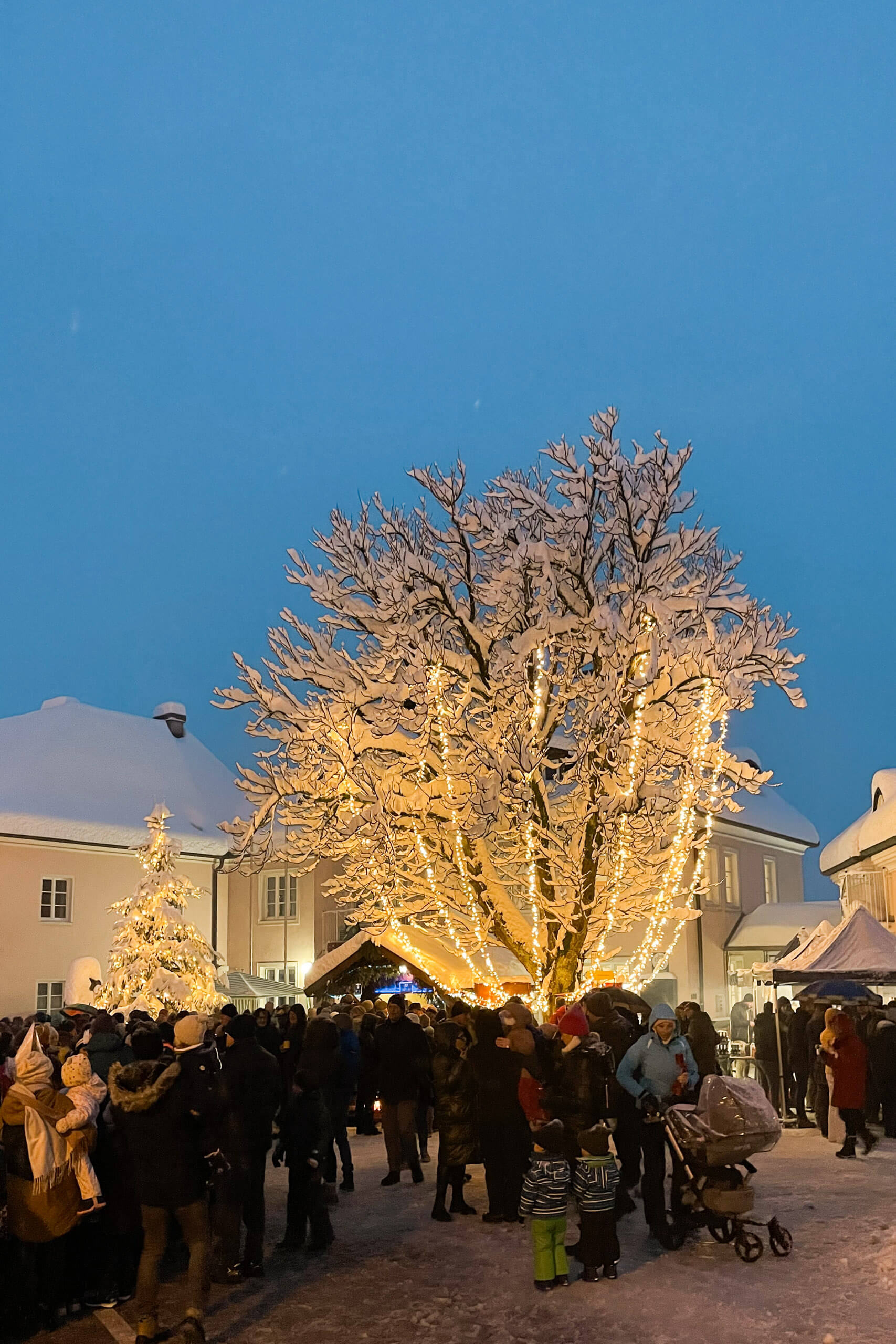 Verschneiter Weihnachtsmarkt mit beleuchtetem Baum und Menschenmenge