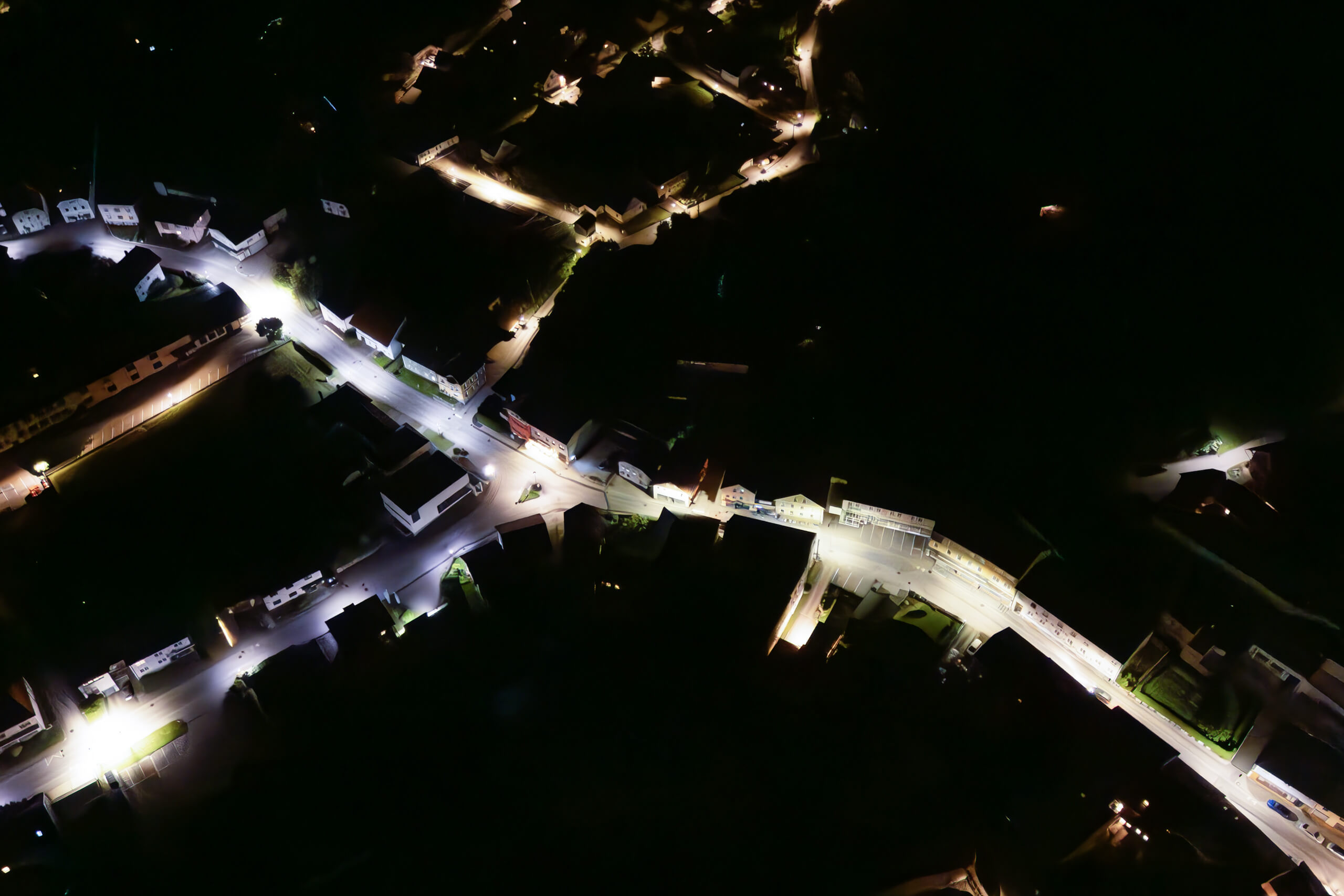 Luftaufnahme einer beleuchteten Ortschaft bei Nacht, Straßenbeleuchtung und Hauslichter sind sichtbar.