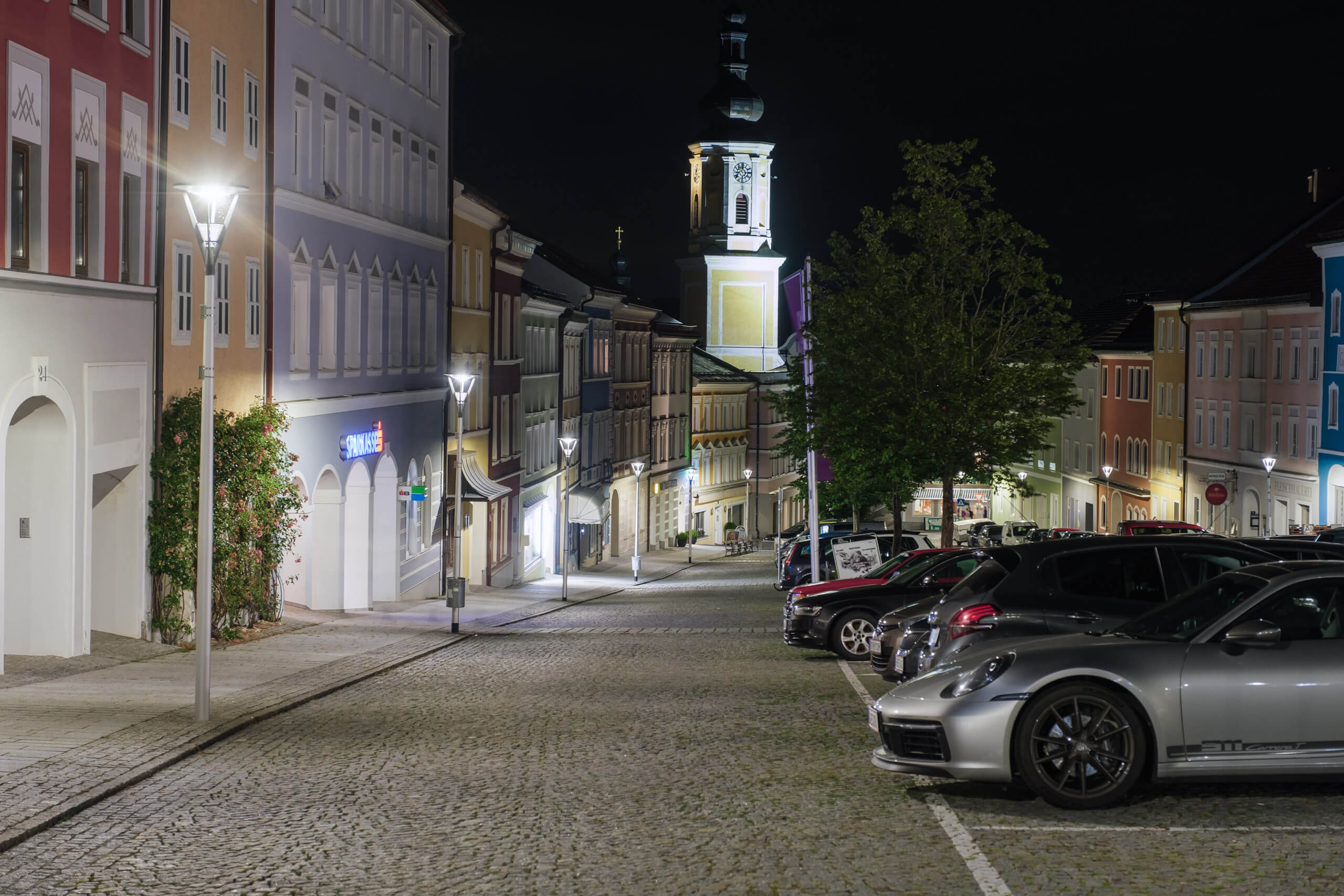 Beleuchtete Stadtstraße mit geparkten Autos und Kirchturm bei Nacht
