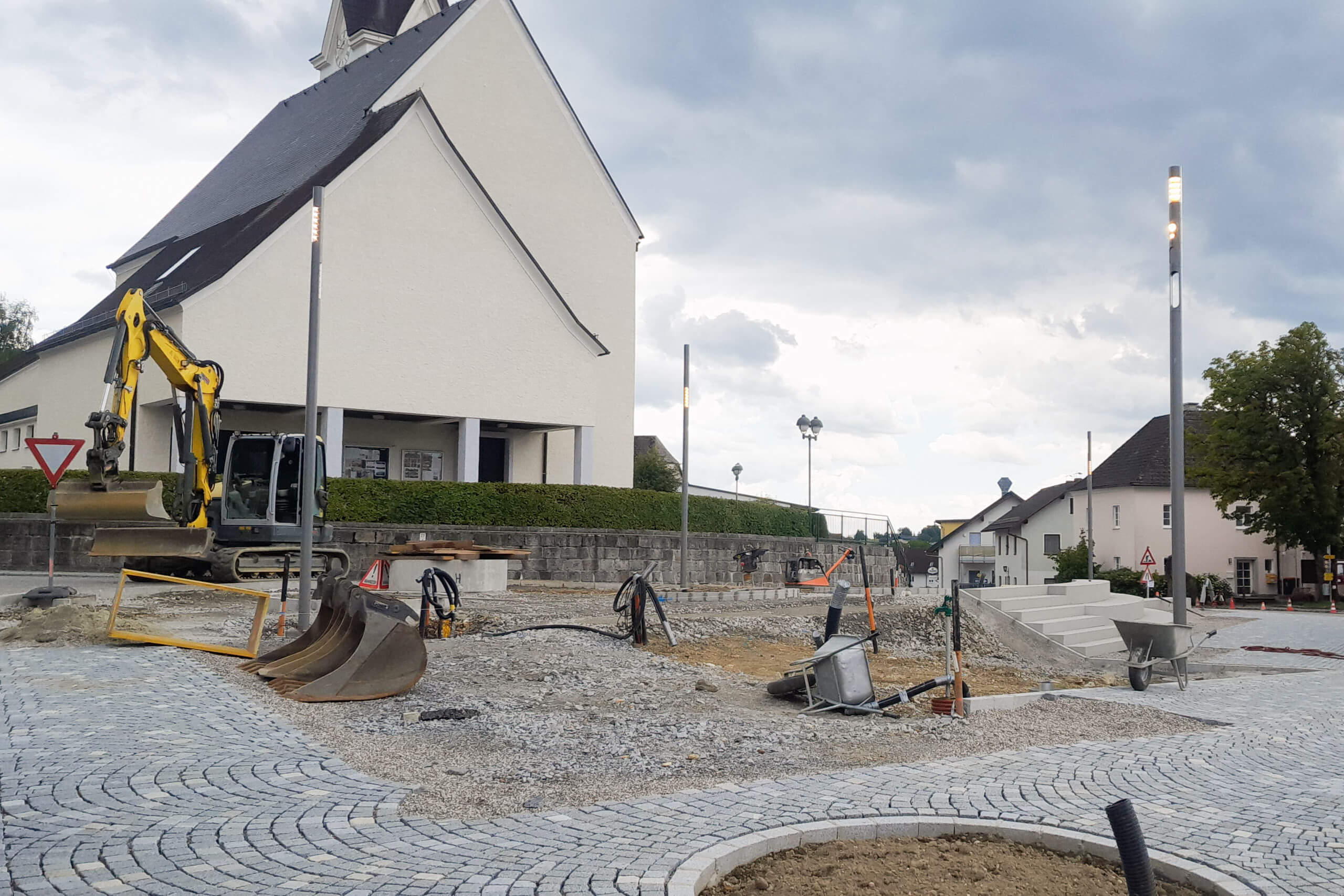 Baustelle mit Bagger vor Kirche, Pflasterarbeiten und moderner Straßenbeleuchtung.