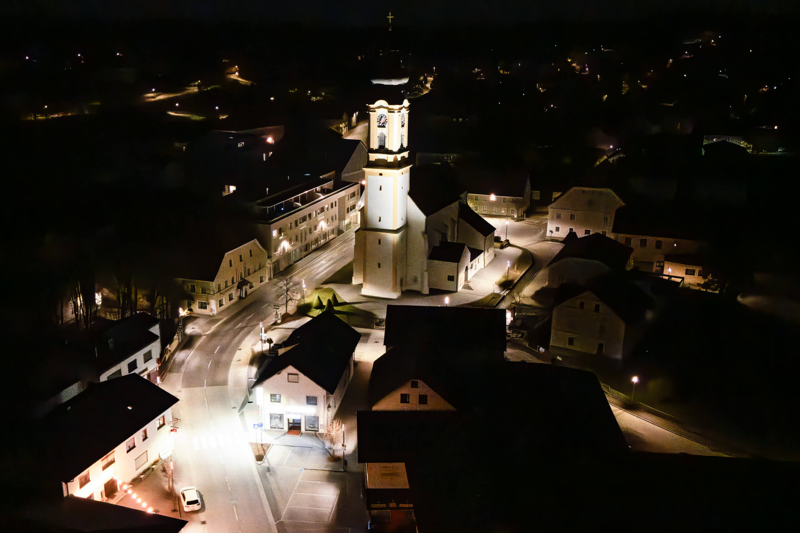 Nachtaufnahme eines beleuchteten Kirchturms und Dorfplatzes mit Straßenbeleuchtung
