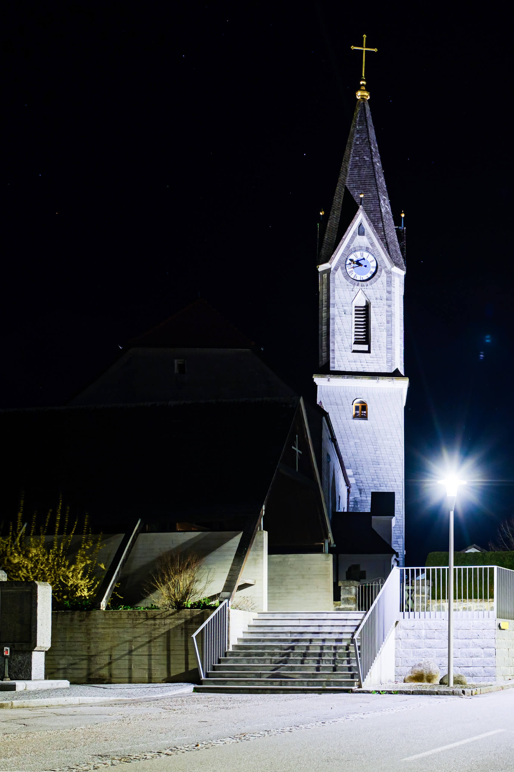 Kirchturm mit Uhr und Kreuz im Nachthimmel, beleuchtet von einer Laterne.