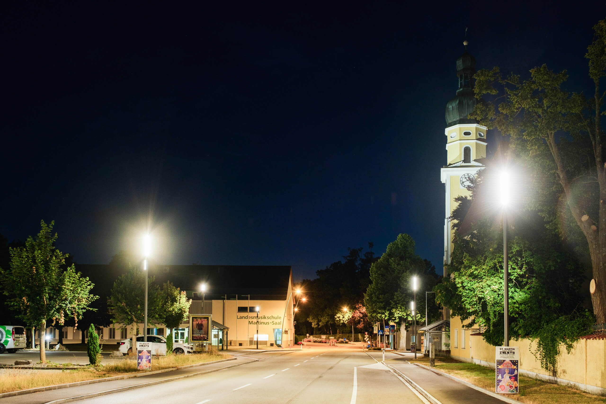Nachtaufnahme einer Straße mit beleuchtetem Kirchturm und Musikschule in Österreich