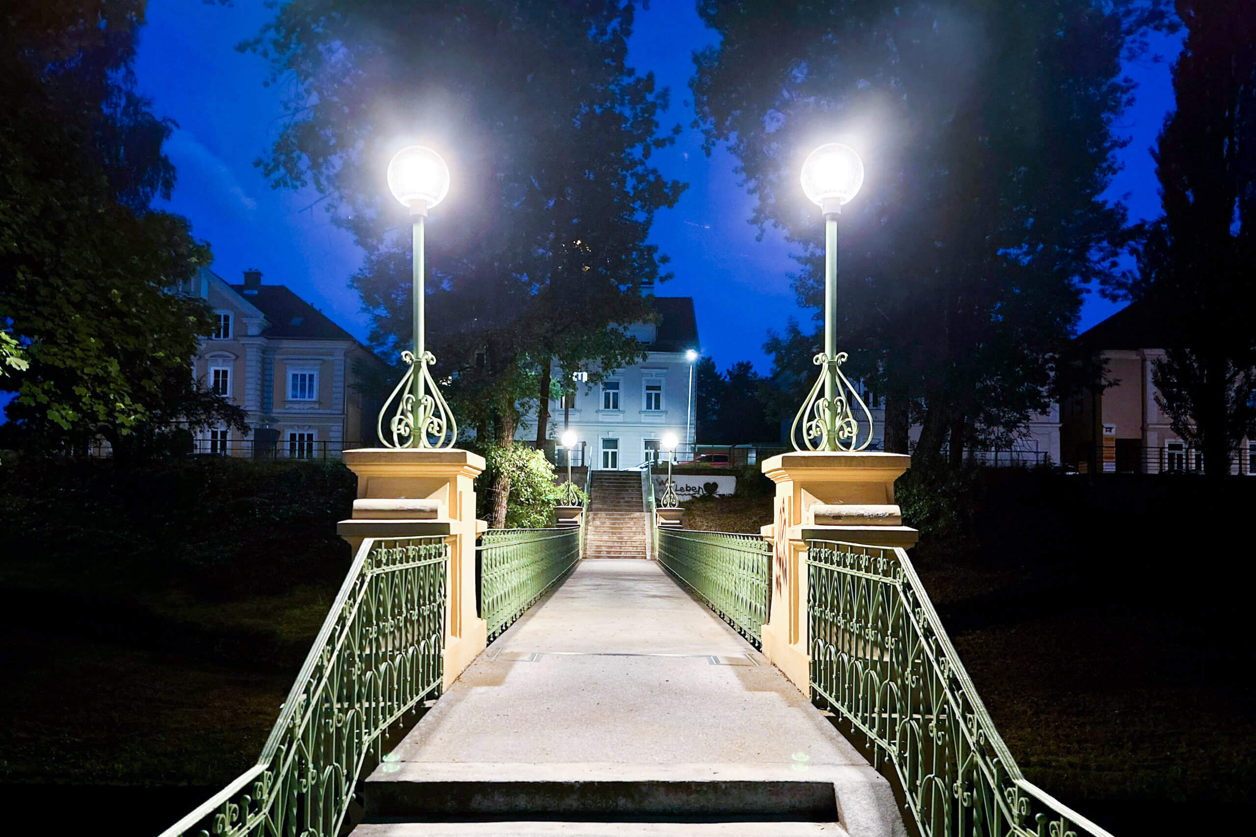 Illumina: Eine beleuchtete Brücke mit Laternen führt zu einem Haus bei Nacht.