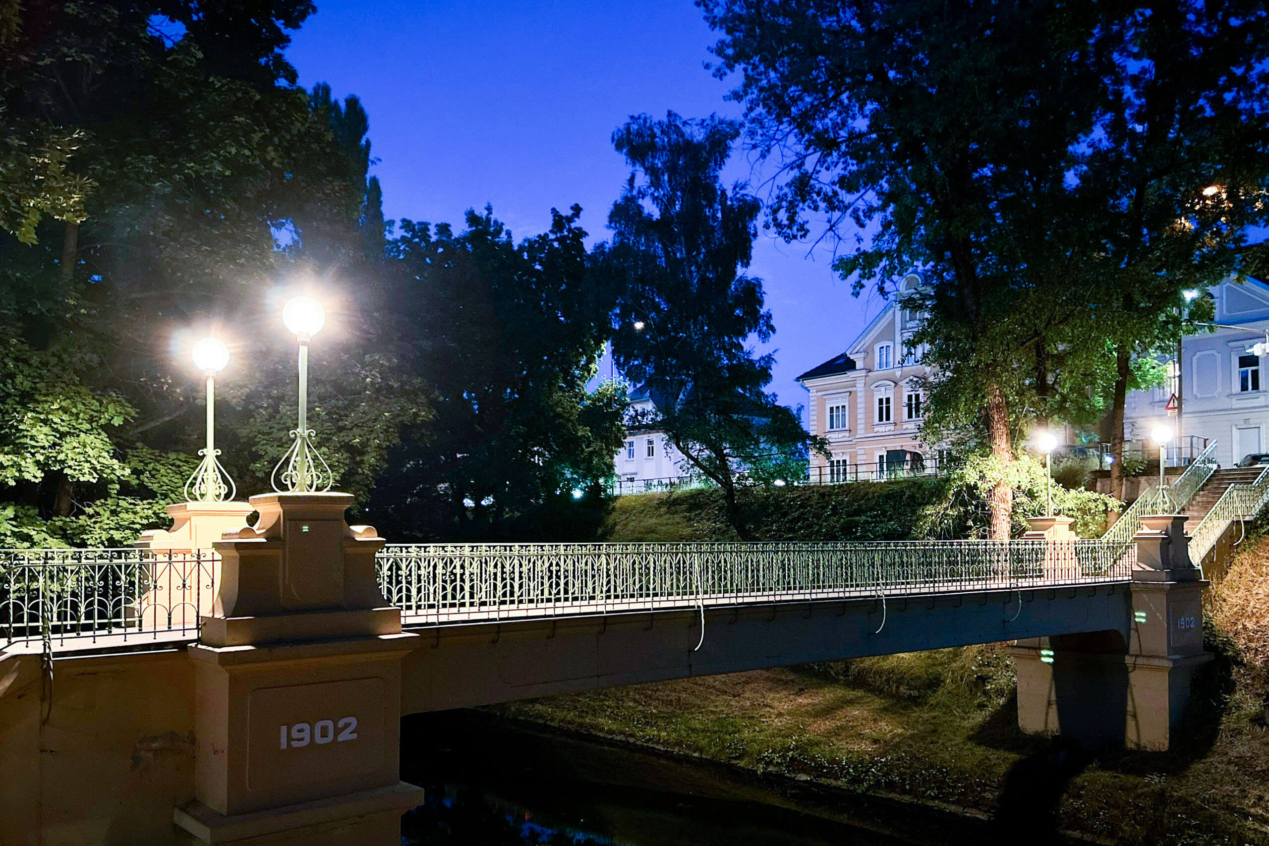 Beleuchtete Brücke aus dem Jahr 1902 mit Laternen und Bäumen im Abendlicht