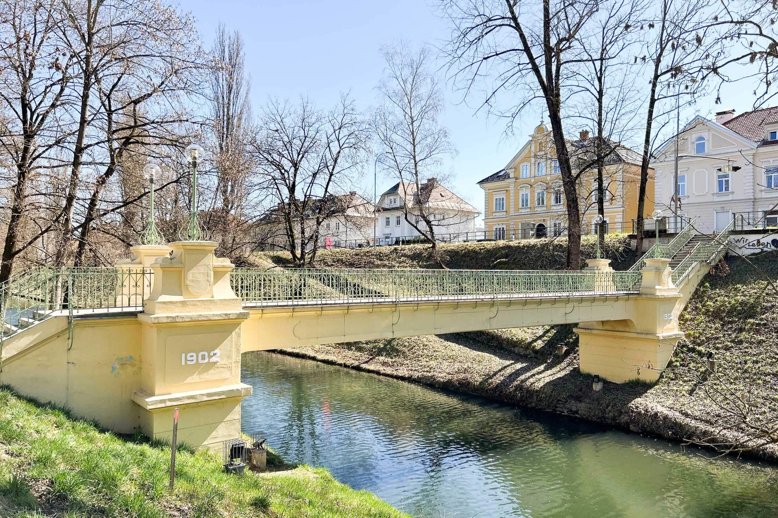 Gelbe Brücke mit der Jahreszahl 1902 über einen Fluss, flankiert von Bäumen und historischen Gebäuden.