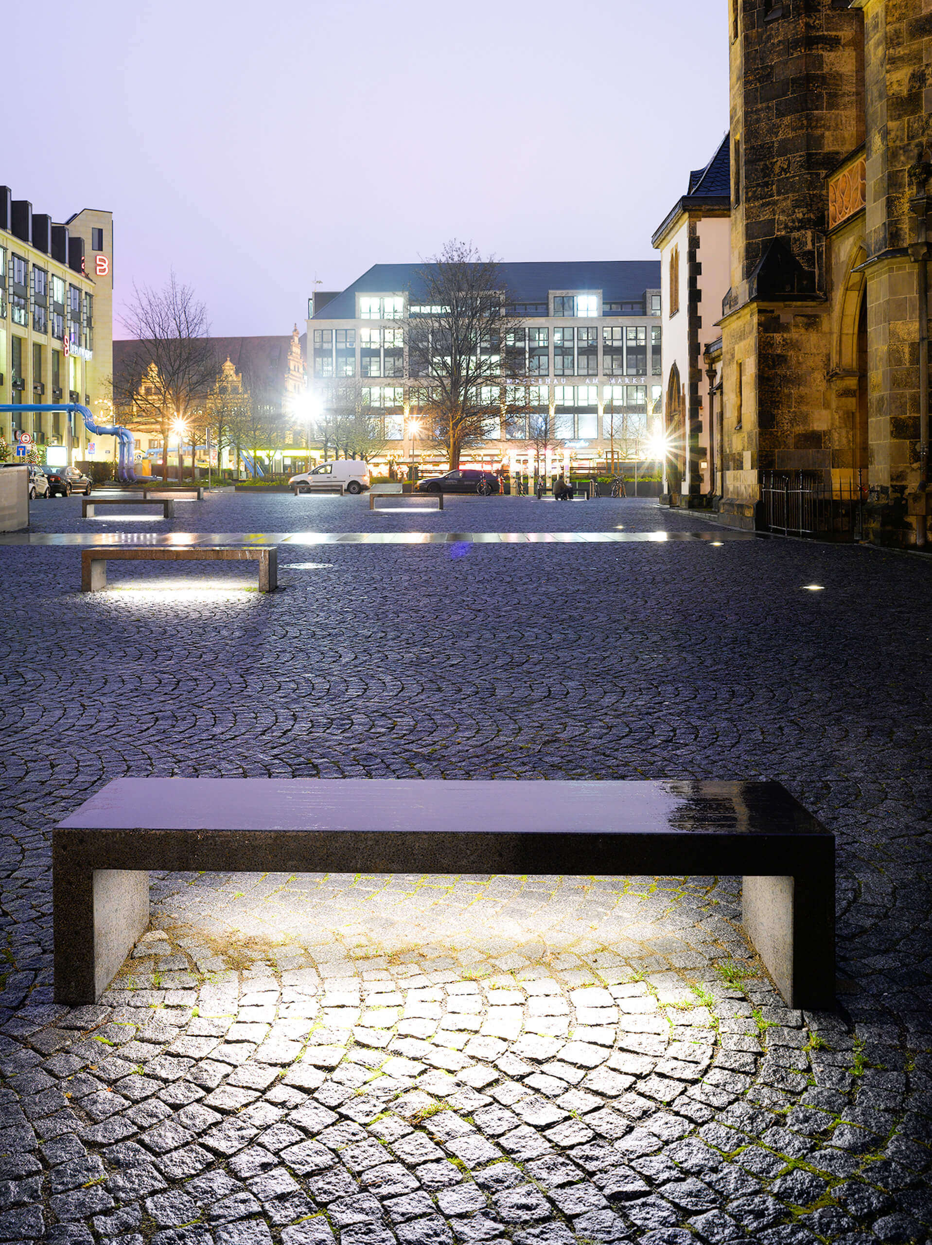 Moderne Stadtmöbel mit integrierter Beleuchtung auf einem Kopfsteinpflasterplatz bei Dämmerung.