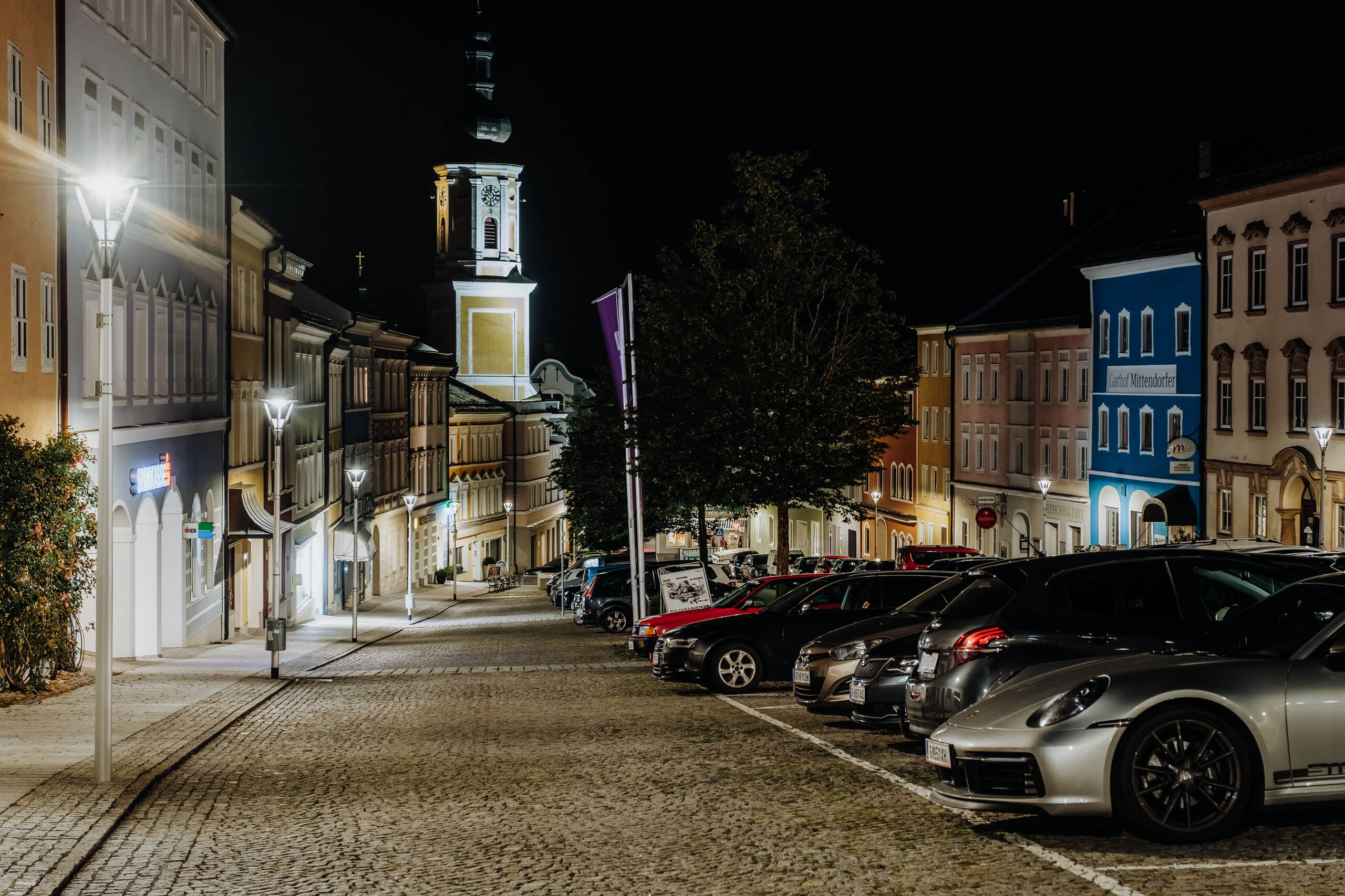 Beleuchtete Altstadtgasse mit geparkten Autos und Kirchturm, Architektur- und Objektanstrahlung