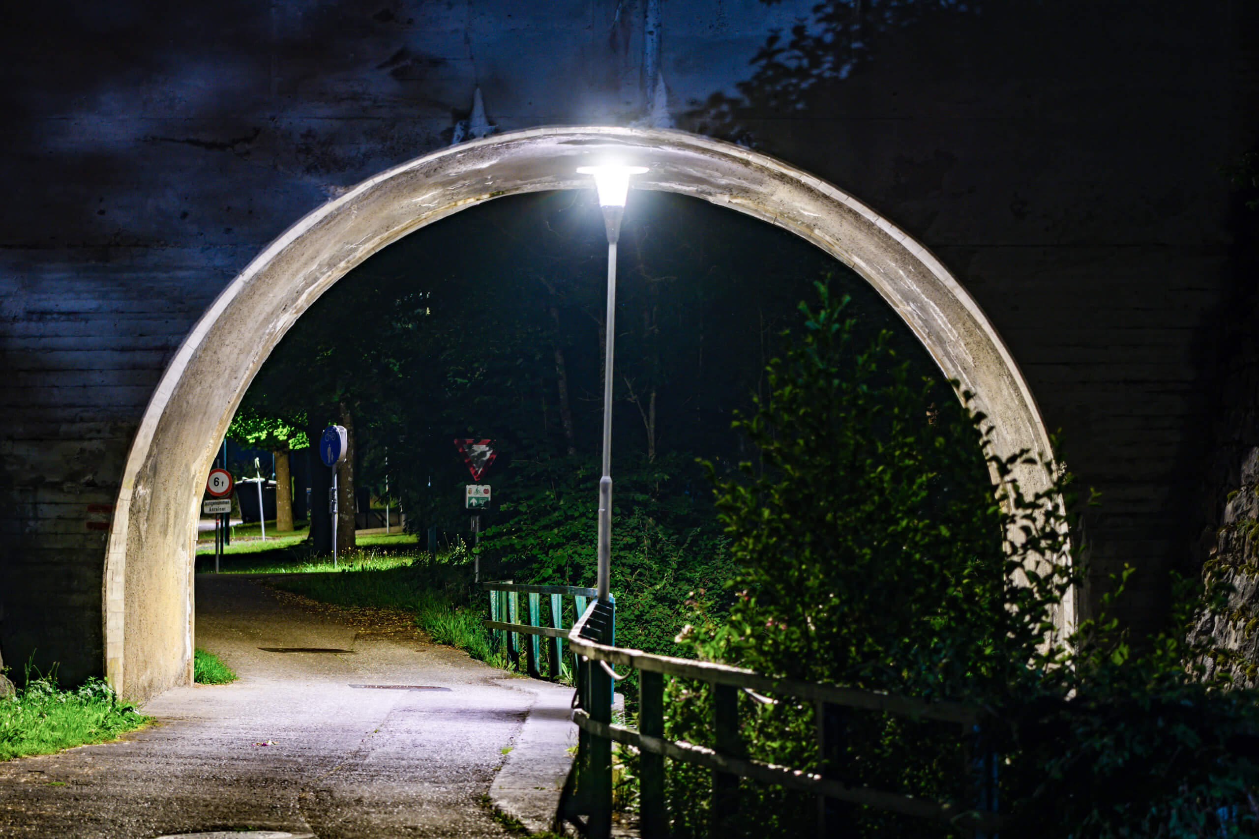 Beleuchteter Fußweg unter einer Betonbrücke mit Vegetation und Straßenschildern