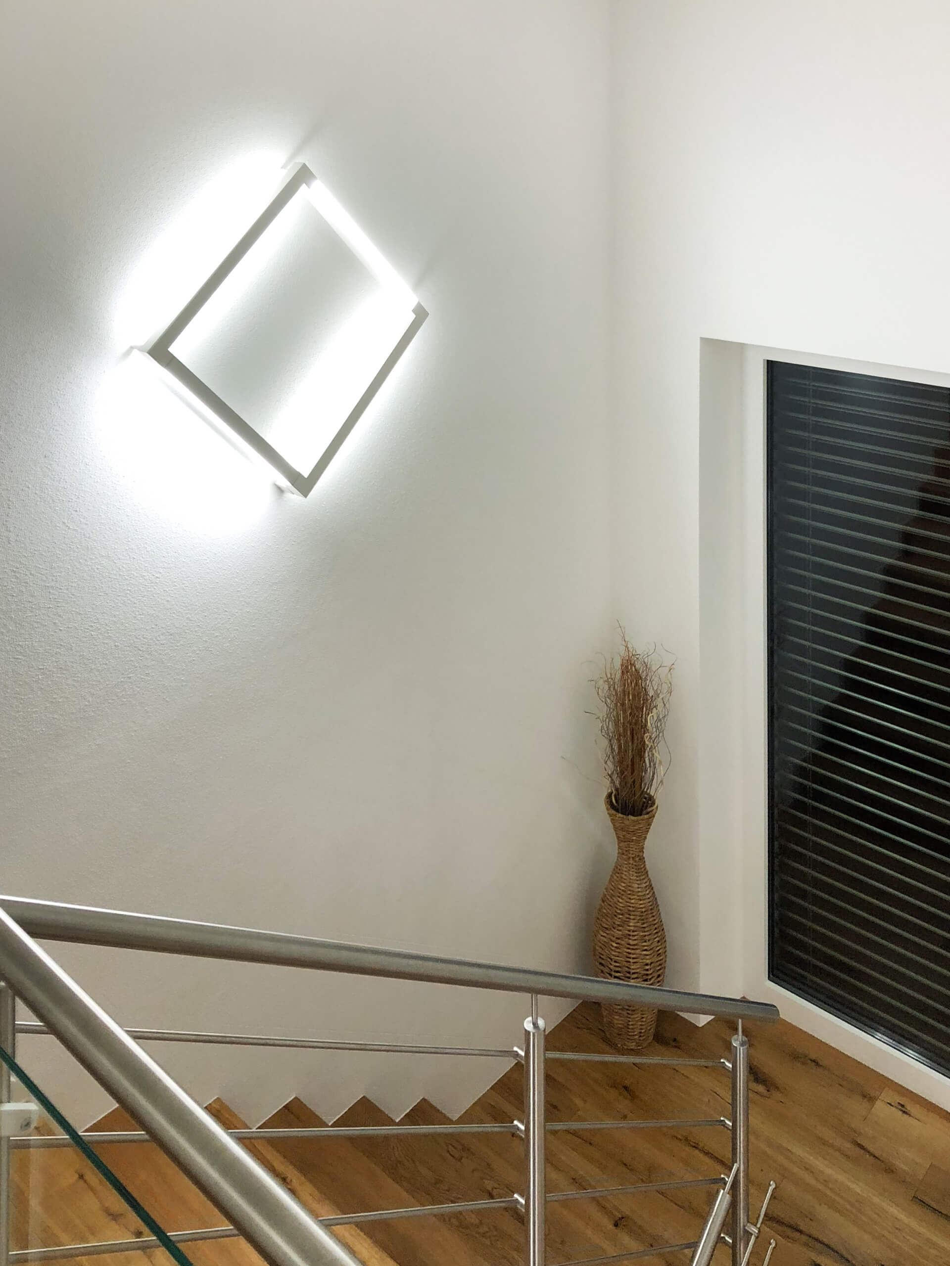 Moderne Treppe mit Glasgeländer und indirekter Beleuchtung, flankiert von einem großen Fenster mit Lamellen und einer dekorativen Vase mit Trockenblumen.