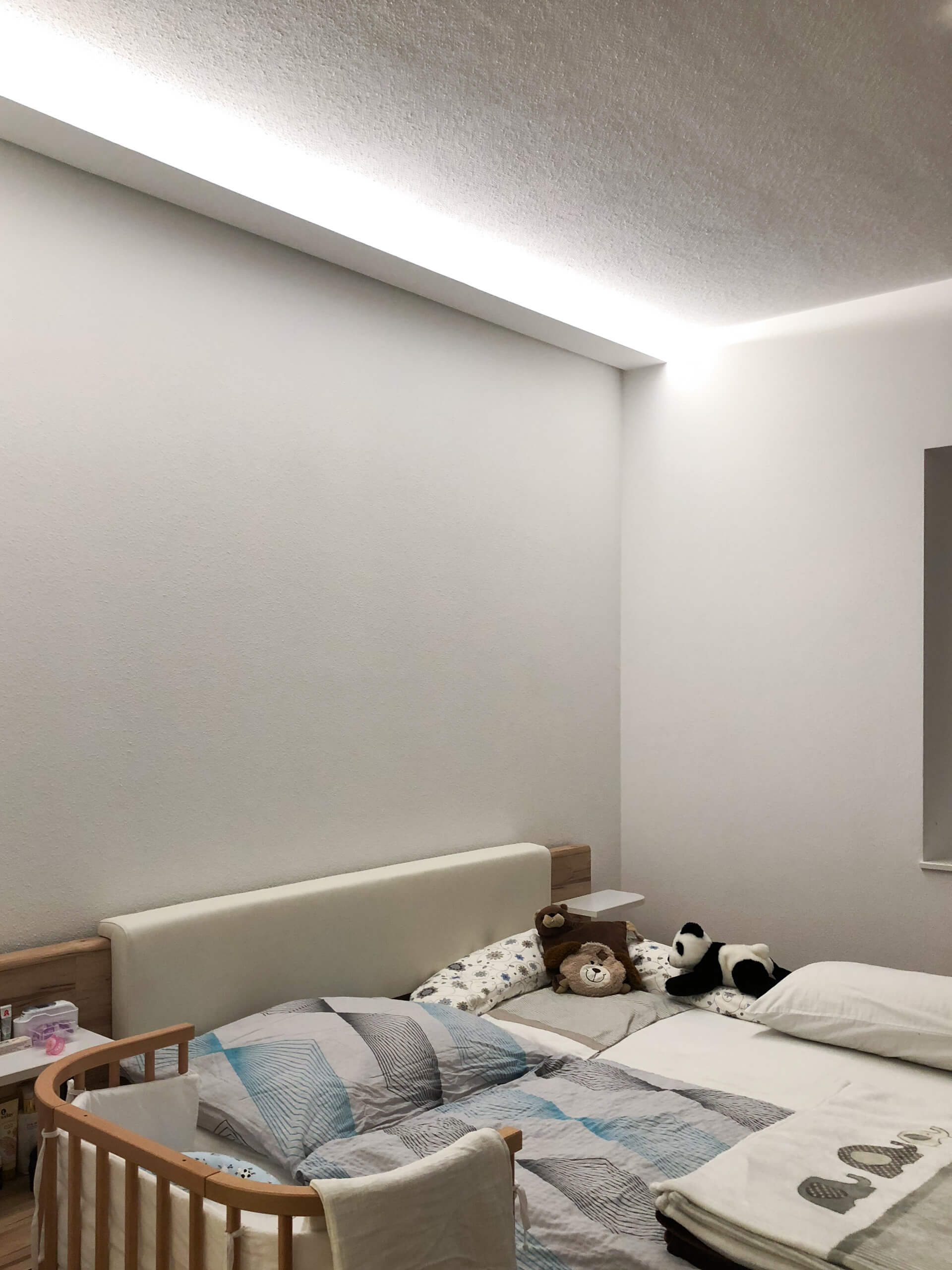 Gemütliches Kinderzimmer mit Bett, Beistellbett und Panda-Spielzeug, beleuchtet durch indirektes Licht.