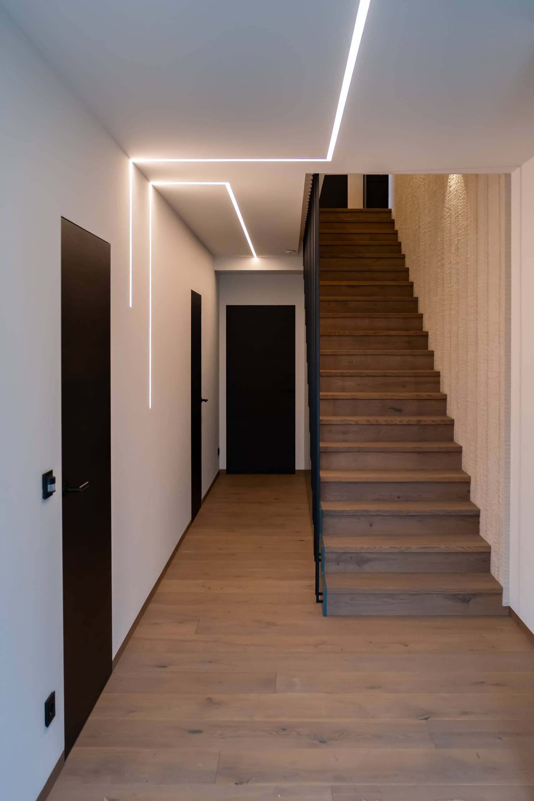 Illuminas Lichtplanung: Moderner Flur mit Holzfußboden, schwarzen Türen und indirekter Deckenbeleuchtung.