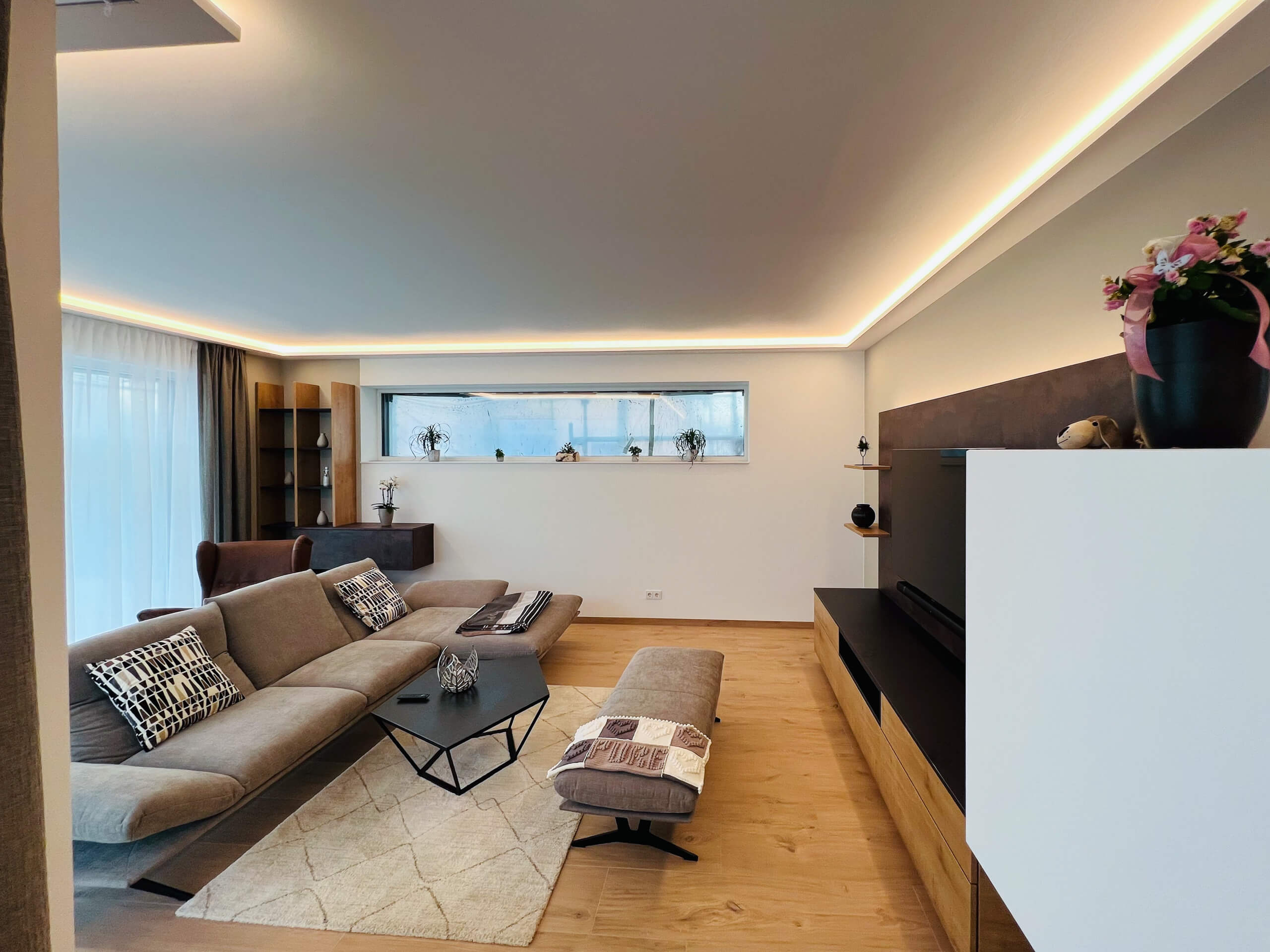 Modernes Wohnzimmer mit indirekter Beleuchtung, beiger Couch und Holzboden.