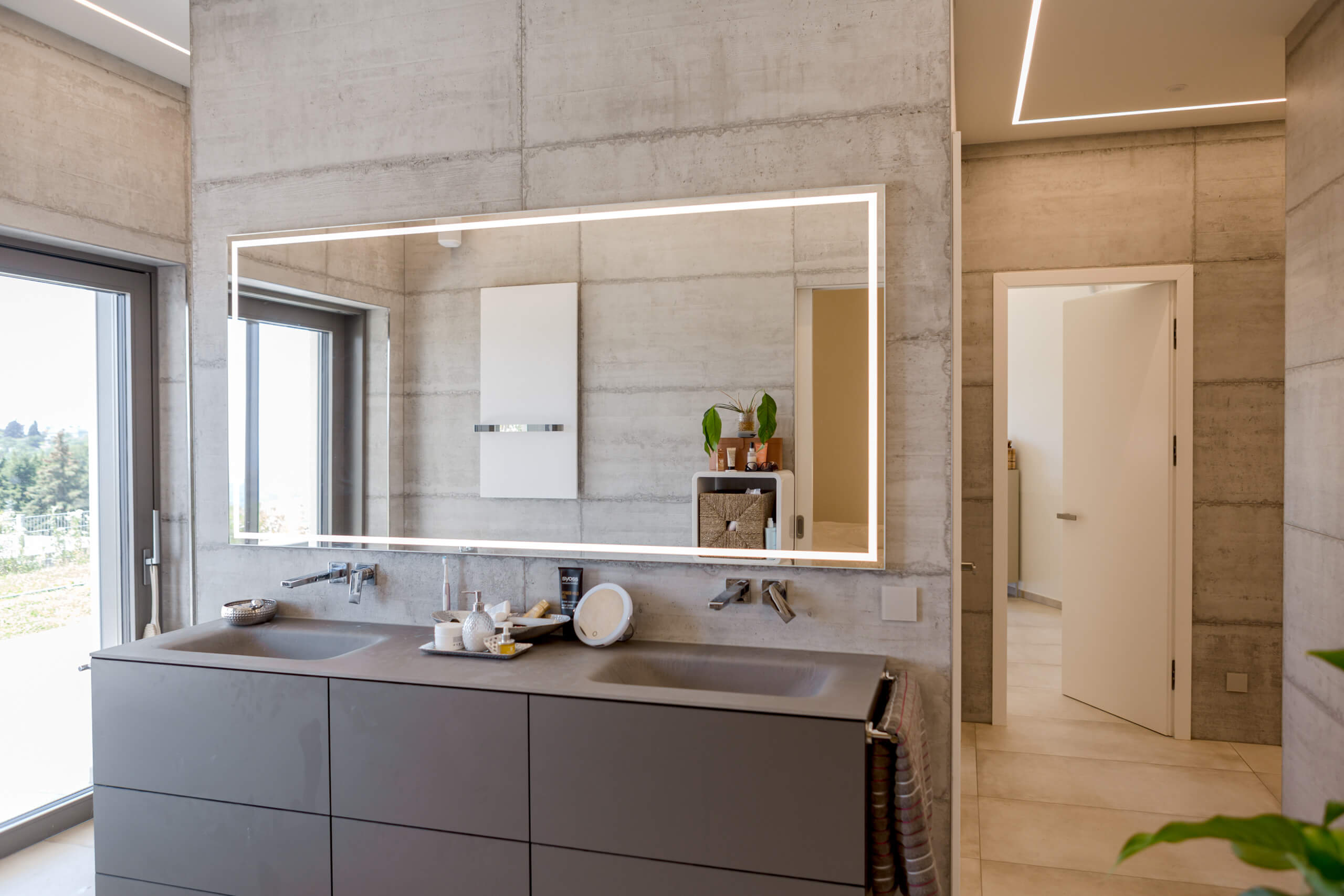 Badezimmer mit Doppelwaschtisch und beleuchtetem Spiegel, Betonwänden und Türe
