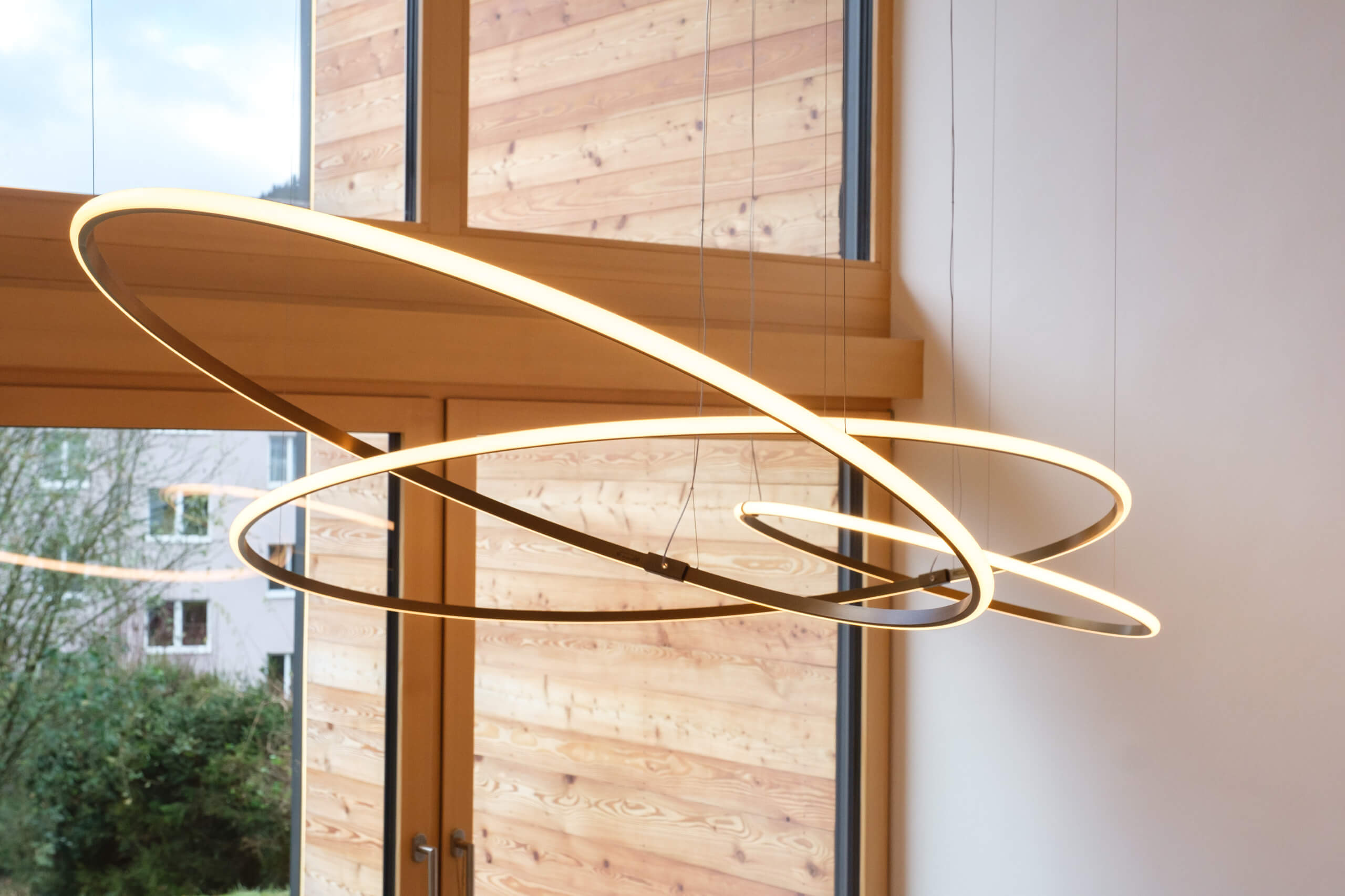 Moderne, geschwungene Hängeleuchte mit warmweißem Licht in einem Raum mit Holzverkleidung.