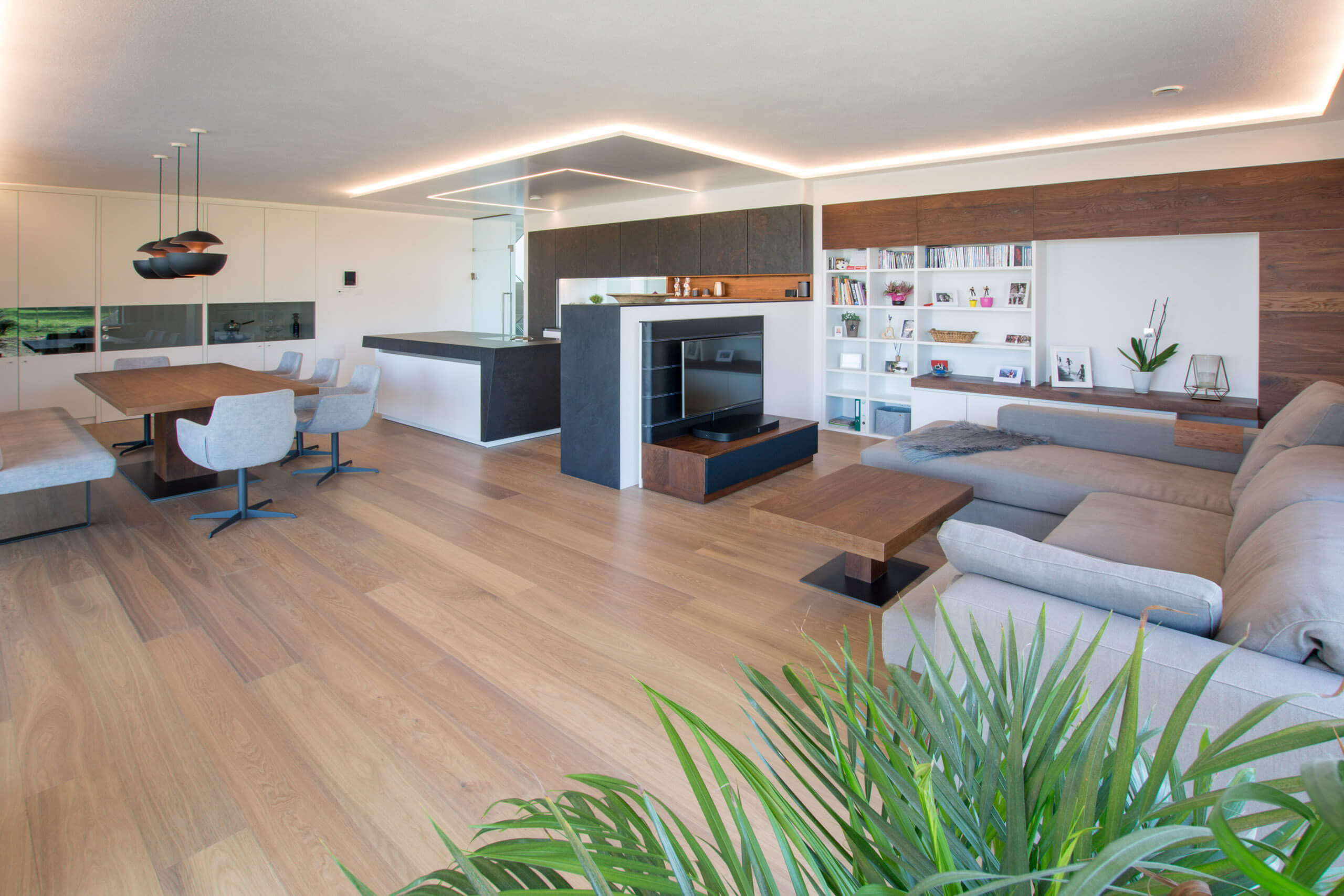 Modernes Wohnzimmer mit integrierter Küche, beleuchtet durch indirektes Licht und Pendelleuchten.