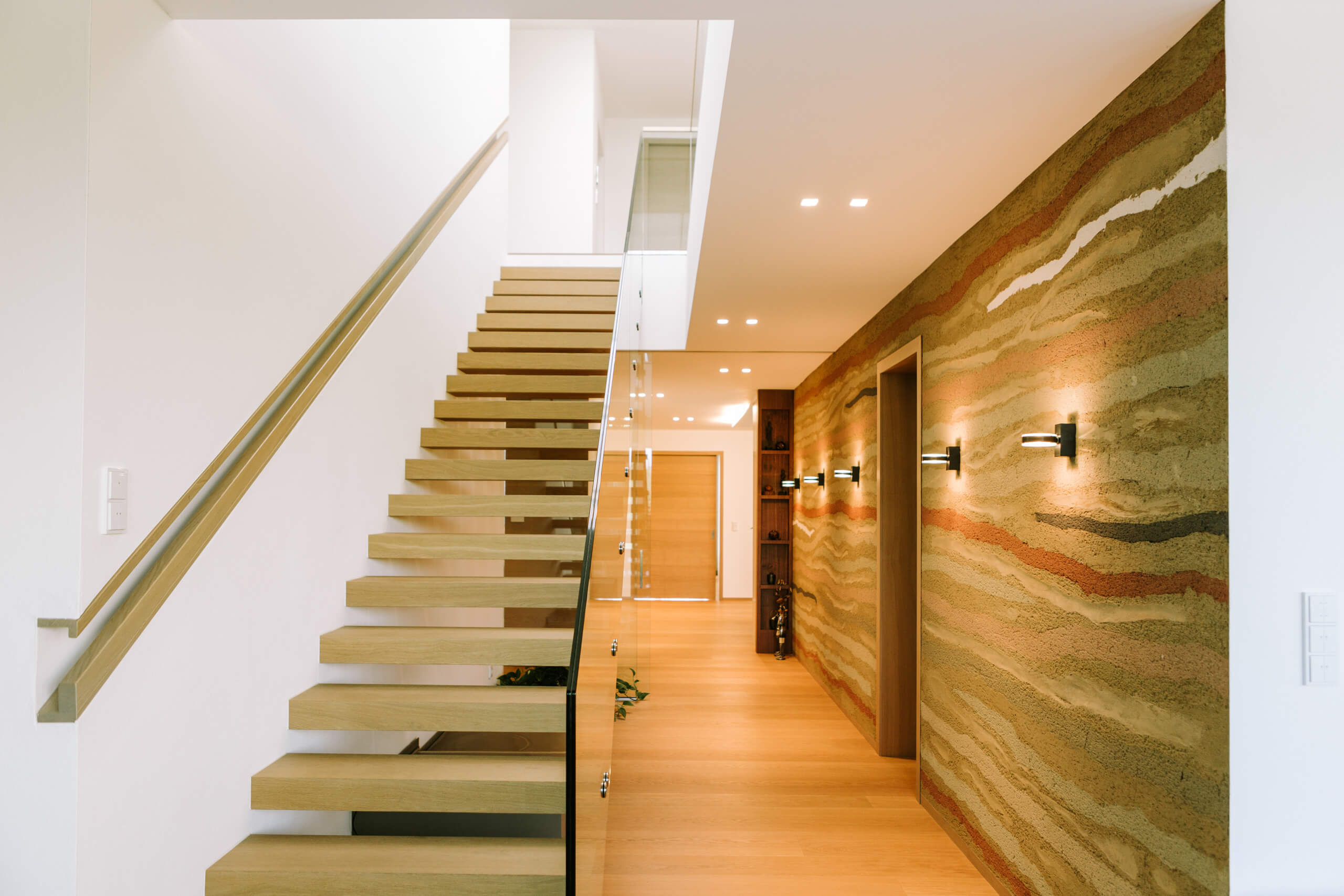 Moderne Holztreppe mit Glasgeländer und erdiger Wandgestaltung – Lichtplanung von Illumina.