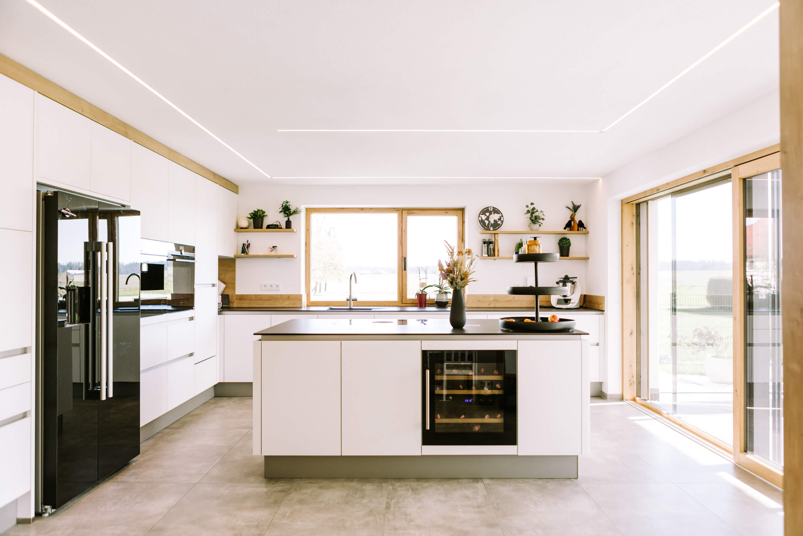 Moderne weiße Küche mit Kochinsel und Wein-Kühlschrank, beleuchtet durch dezente Deckenbeleuchtung.