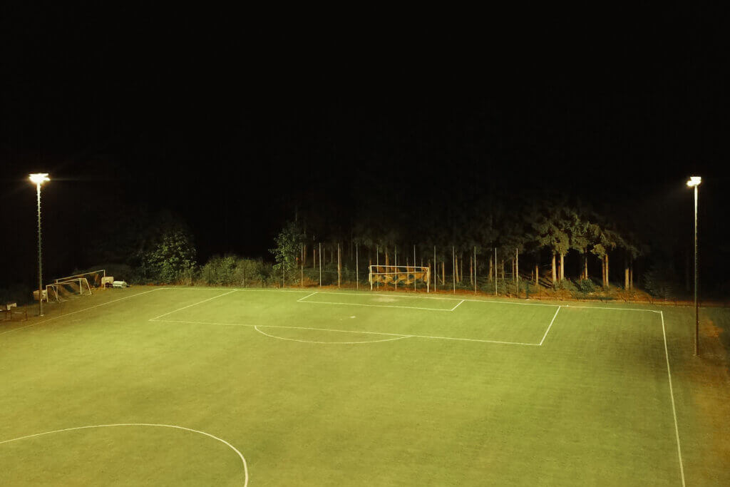 Fußballplatz bei Nacht mit Flutlichtbeleuchtung und Wald im Hintergrund.