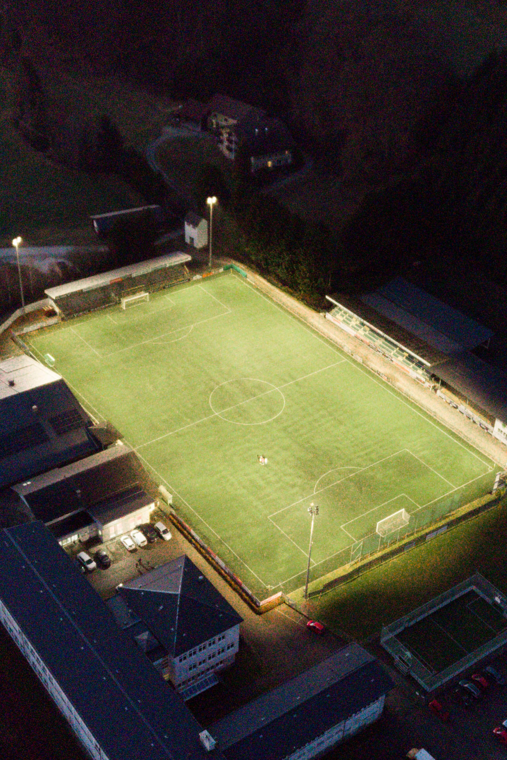 Fußballplatz mit Flutlichtbeleuchtung bei Nacht, umgeben von Gebäuden und dunkler Natur.