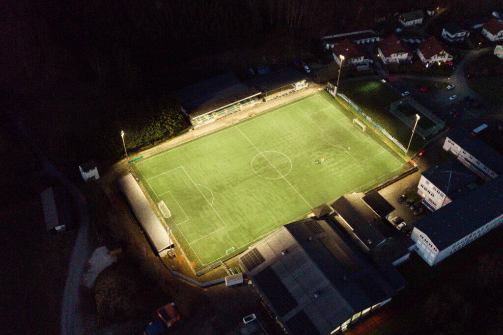 Illuminas Sportplatzbeleuchtung: Ein beleuchteter Fußballplatz bei Nacht mit Tribüne und angrenzenden Gebäuden.