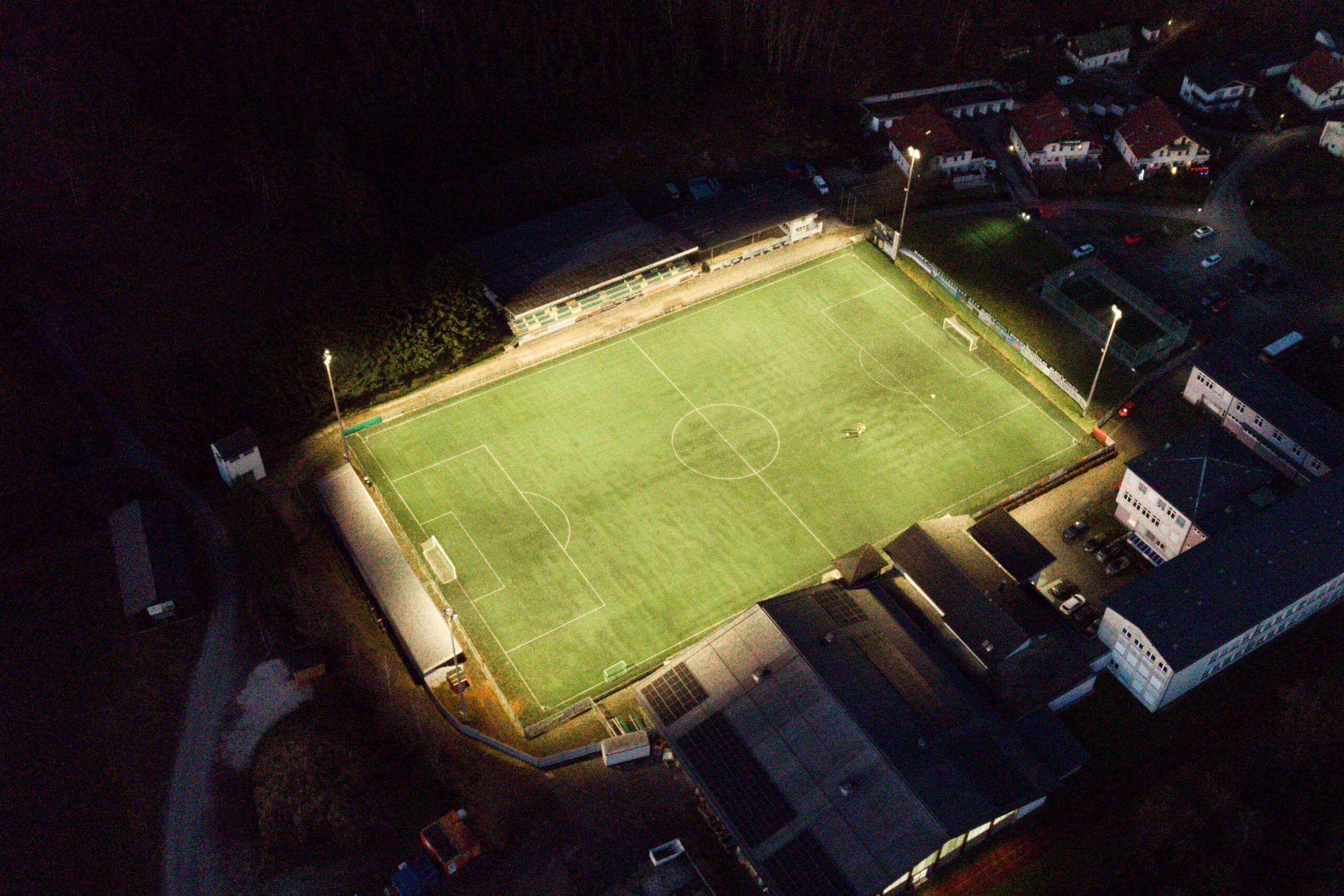 Illuminas Sportplatzbeleuchtung: Ein beleuchteter Fußballplatz bei Nacht mit Tribüne und angrenzenden Gebäuden.