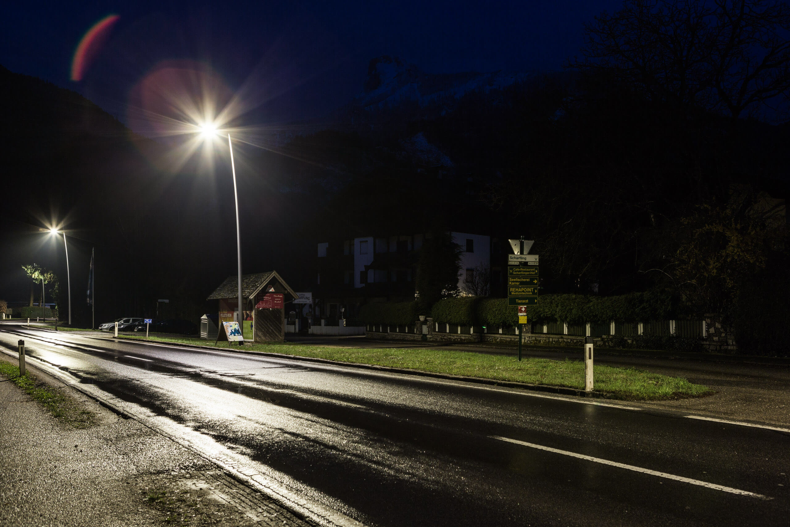 Straßenbeleuchtung und Ortstafel im Dunkeln, beleuchtet von Laternen, mit Berg im Hintergrund.