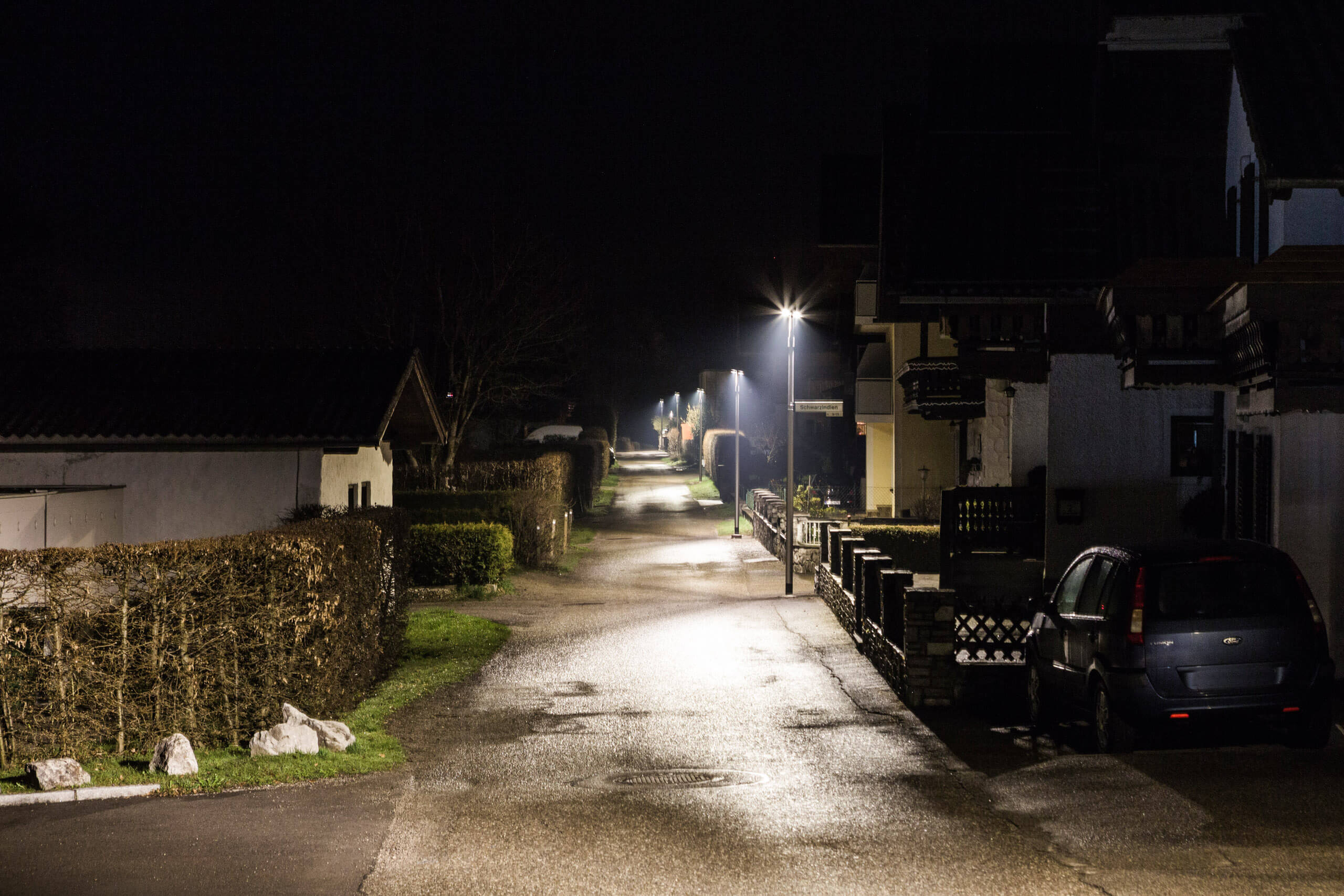 Illumina: Straßenbeleuchtung in einer dunklen Gasse mit gepflegten Hecken und Häusern.