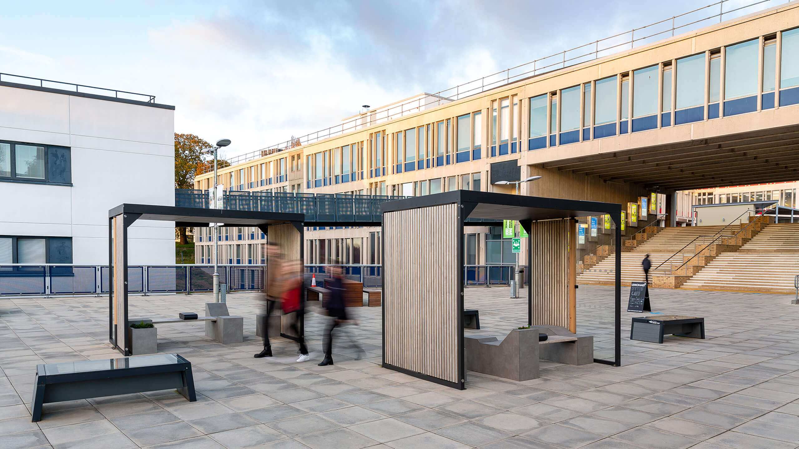 Moderne Stadtmobiliar-Elemente mit Sitzbänken und Überdachungen auf einem gepflasterten Platz.