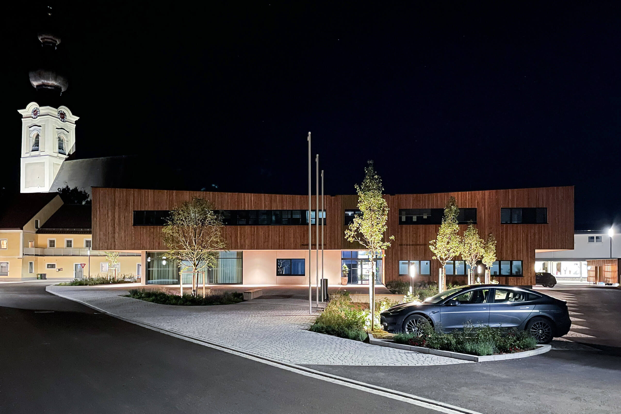 Moderne Holzarchitektur mit Fassadenbeleuchtung und Tesla vor Kirche bei Nacht