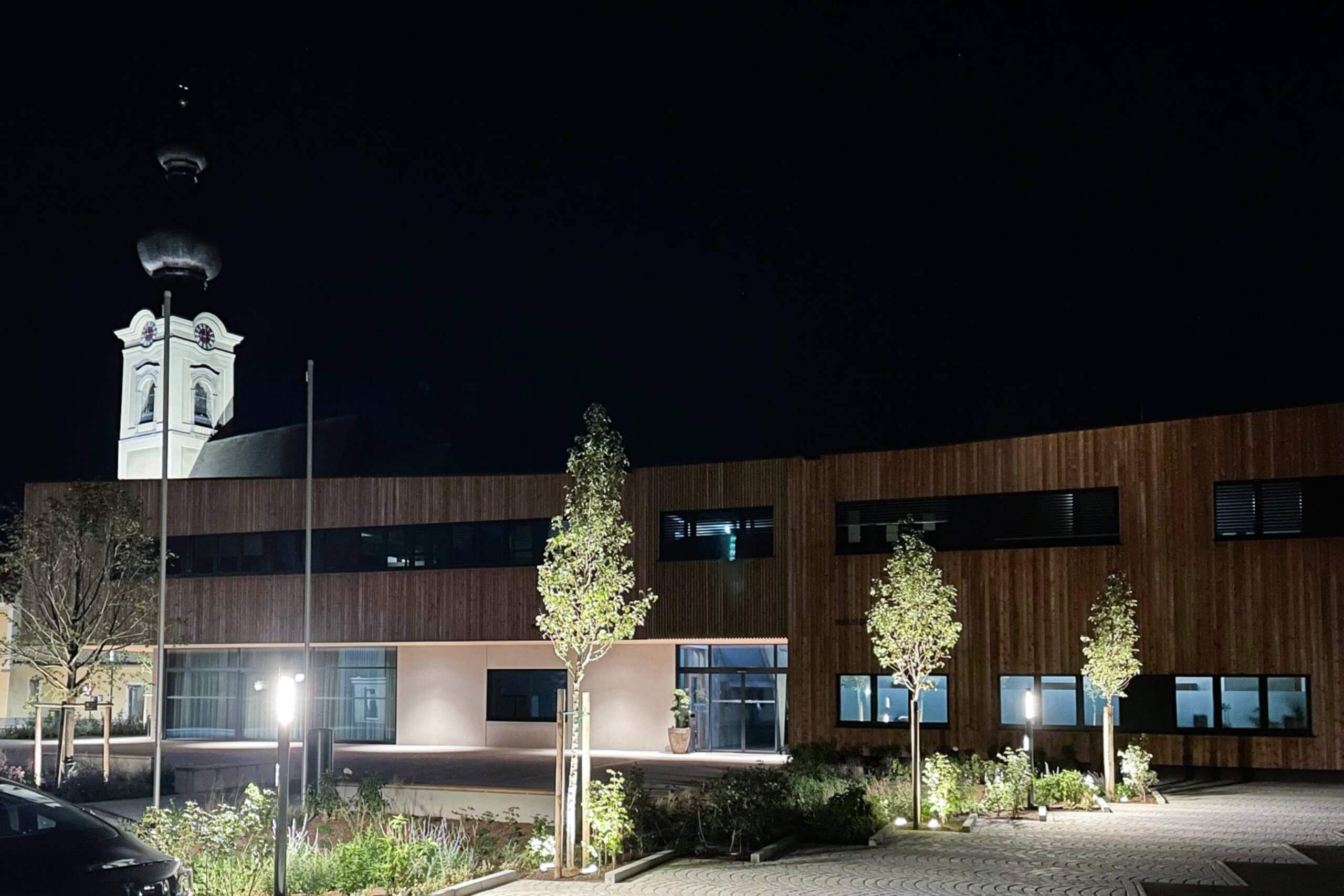 Moderne Holzfassade mit Architekturbeleuchtung und Kirchturm bei Nacht